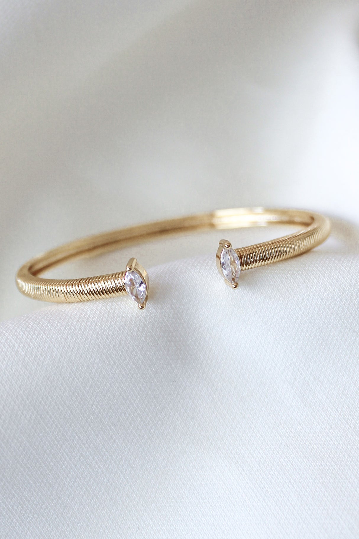The Mara Cuff by Kinsey Designs is a gold plated adjustable bracelet, showcasing two clear cubic zirconia gemstones on each end, displayed on a white fabric background.