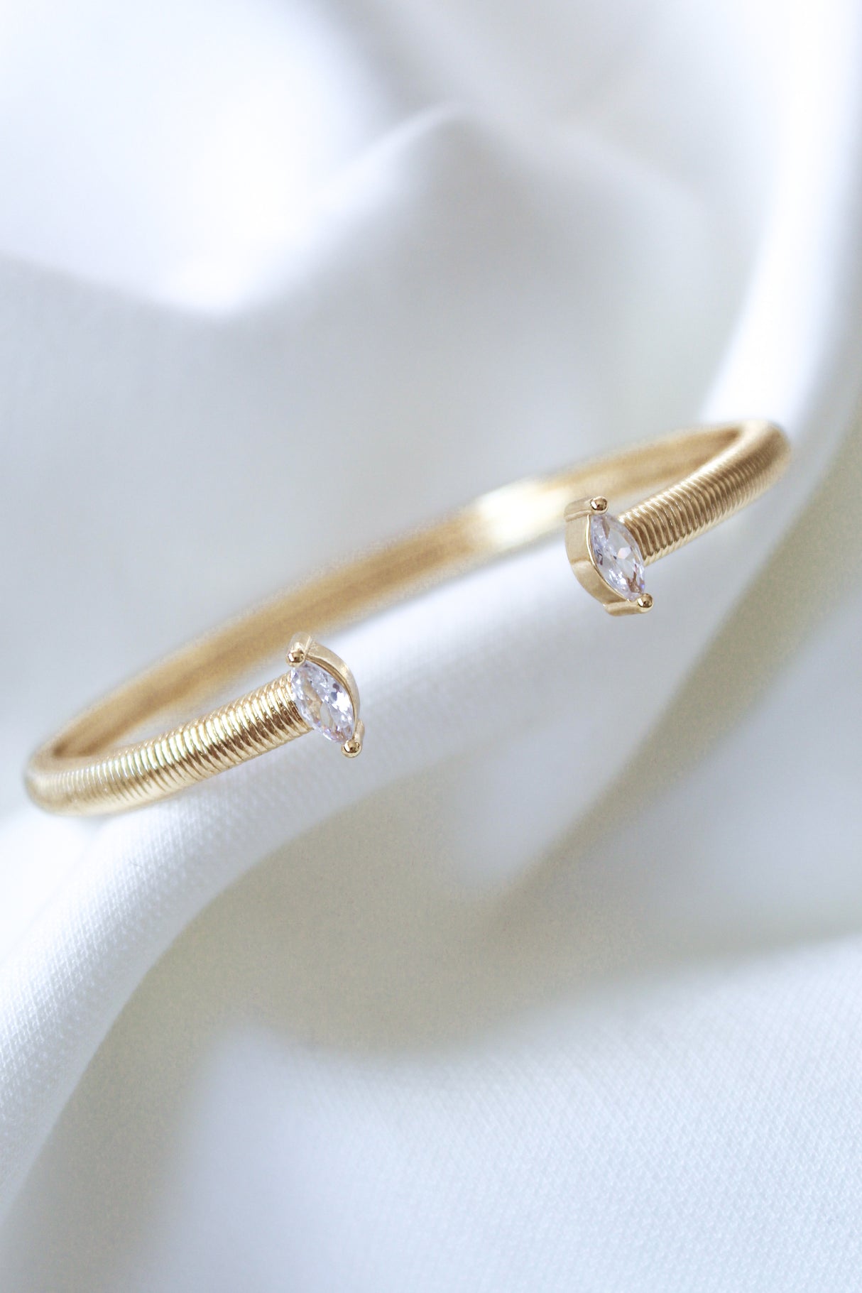 The Mara Cuff by Kinsey Designs is a gold-plated bracelet featuring two marquise-cut cubic zirconia stones, elegantly displayed on soft white fabric and adjustable for a perfect fit.
