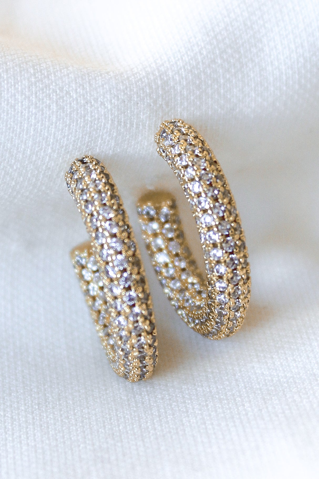 The Melli Hoop earrings by Kinsey Designs feature gold-filled hoops adorned with small sparkling cubic zirconia, shown on a soft white fabric background.