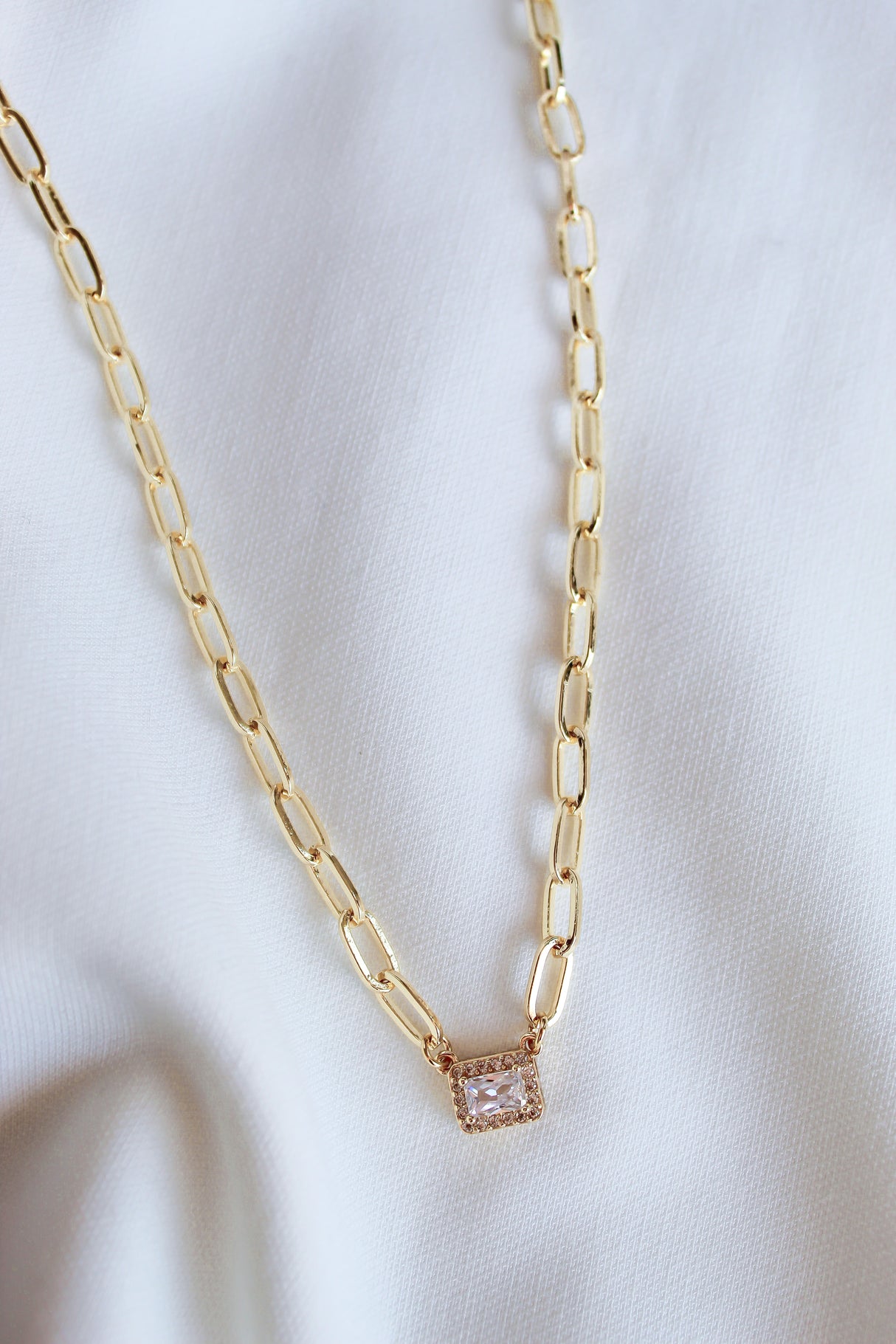 The Posh Necklace by Kinsey Designs features a gold-filled rectangular pendant, elegantly displayed on white fabric—a perfect statement piece for any occasion.