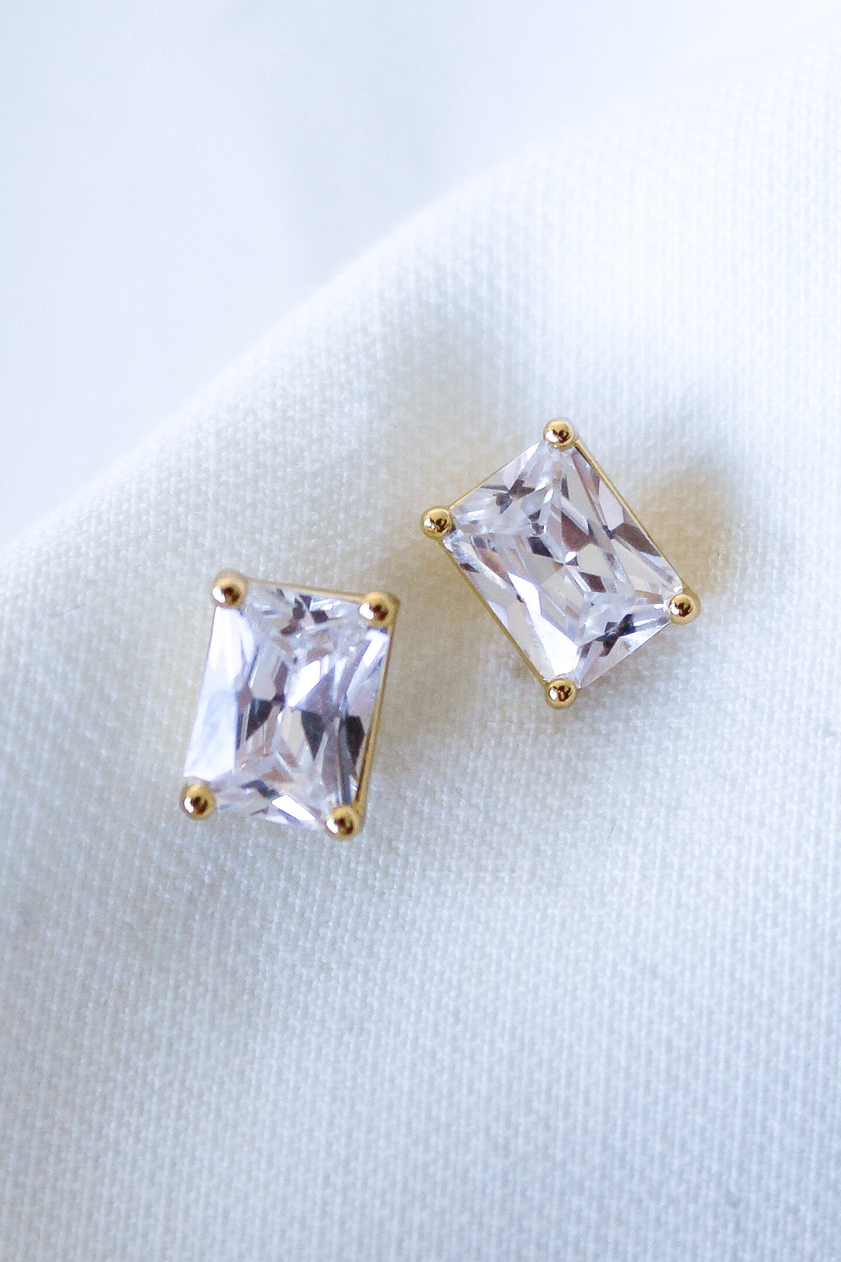 Two rectangular Kinsey Designs Prism Studs with gold prongs rest on white fabric. These elegant Cubic Zirconia earrings offer a modern update to classic style.