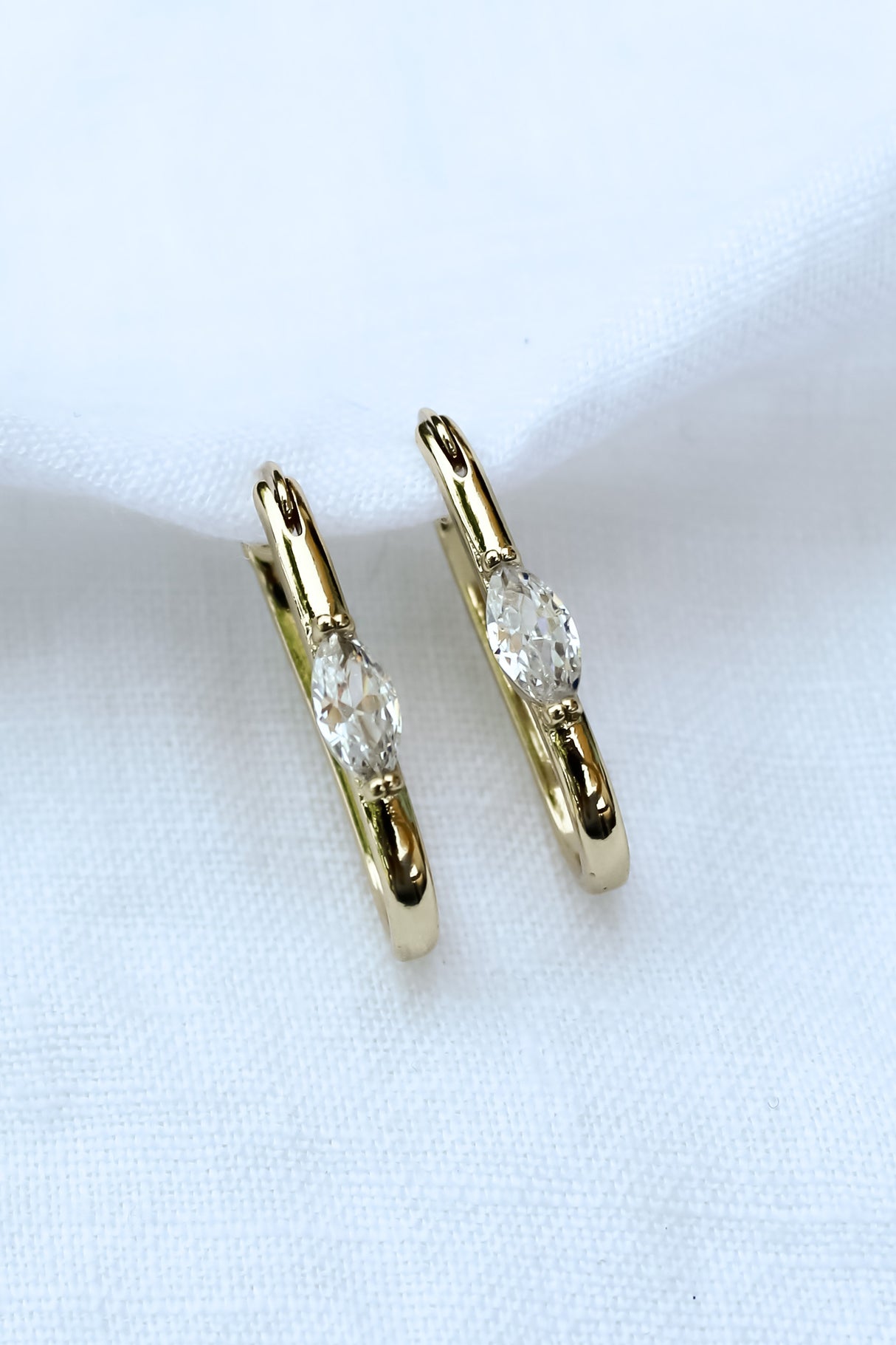 The Ragan Huggie by Kinsey Designs Archives features gold filled hoop earrings with round cubic zirconia gemstones, 1" long, displayed on a white fabric background.