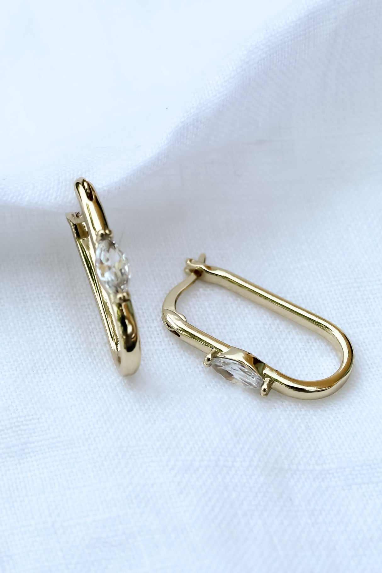 The Ragan Huggie earrings by Kinsey Designs Archives feature gold-filled hoops, 1" long, with sparkling cubic zirconia gemstones, shown on a white fabric background.