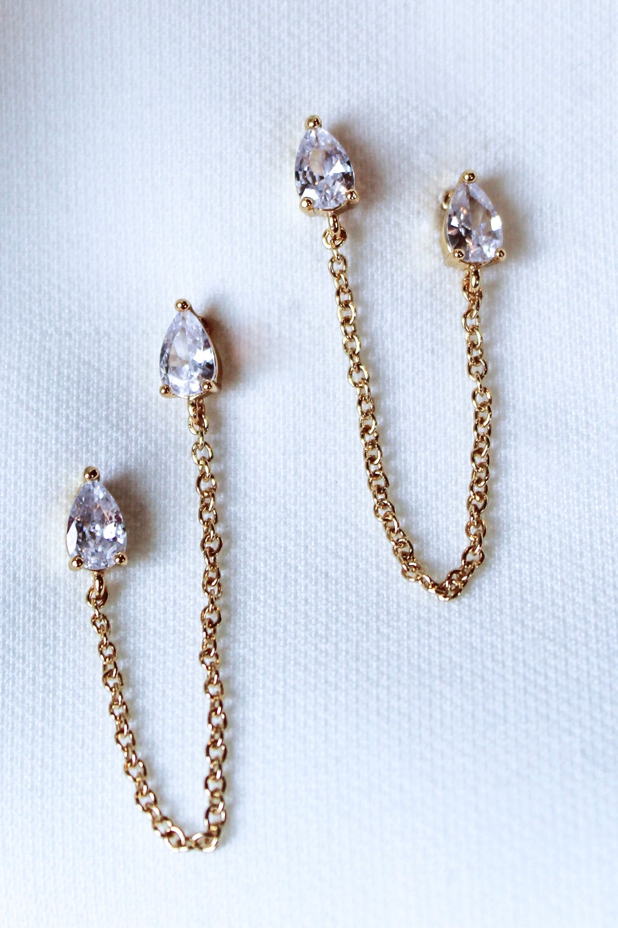 The Rayna Post earrings by Kinsey Designs Archives, featuring gold-plated chains and teardrop-shaped cubic zirconia, are displayed on a light fabric background.