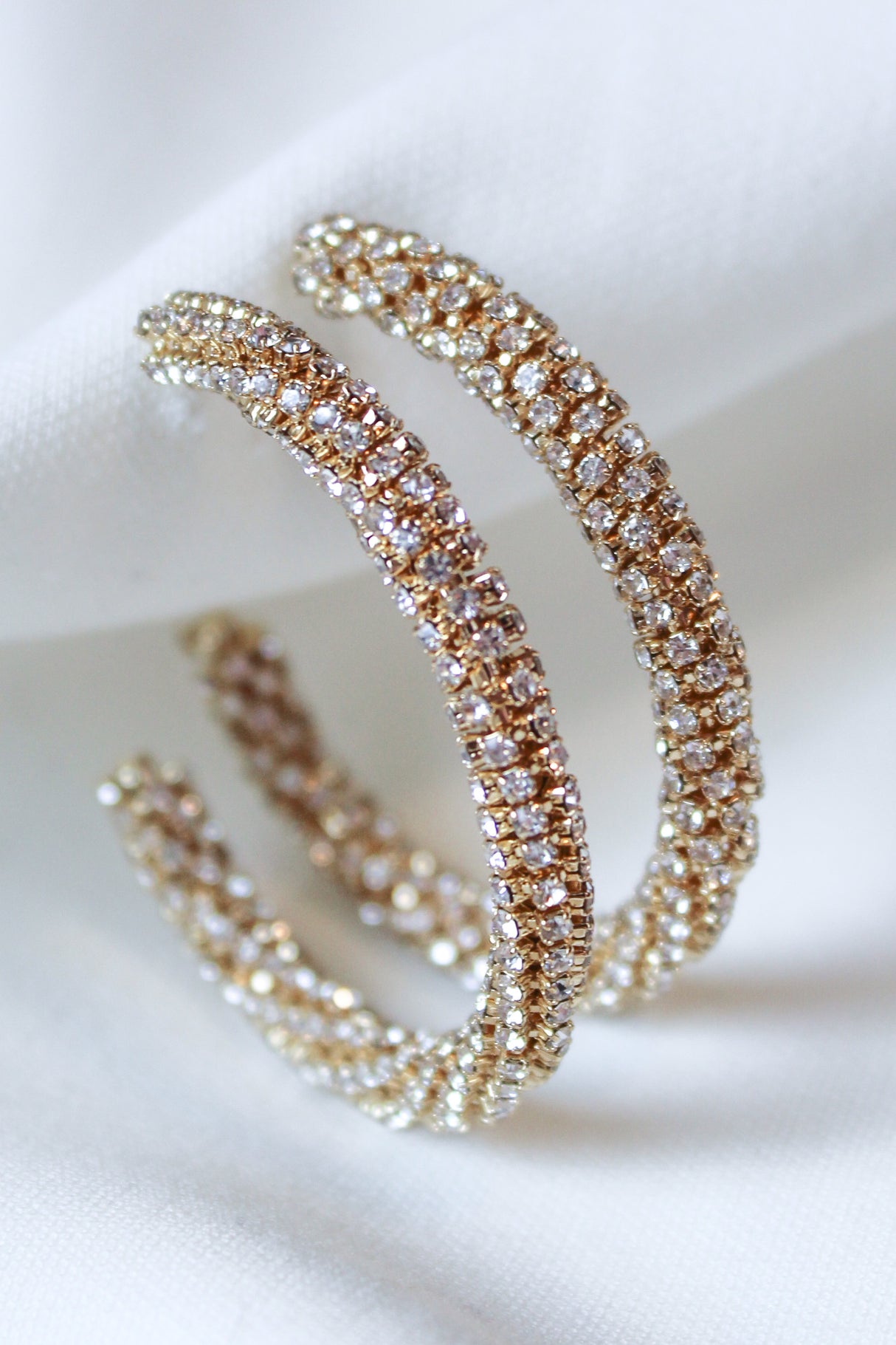 The Riley Hoop earrings by Kinsey Designs feature gold plating and are encrusted with sparkling cubic zirconia, shown on a white fabric background.