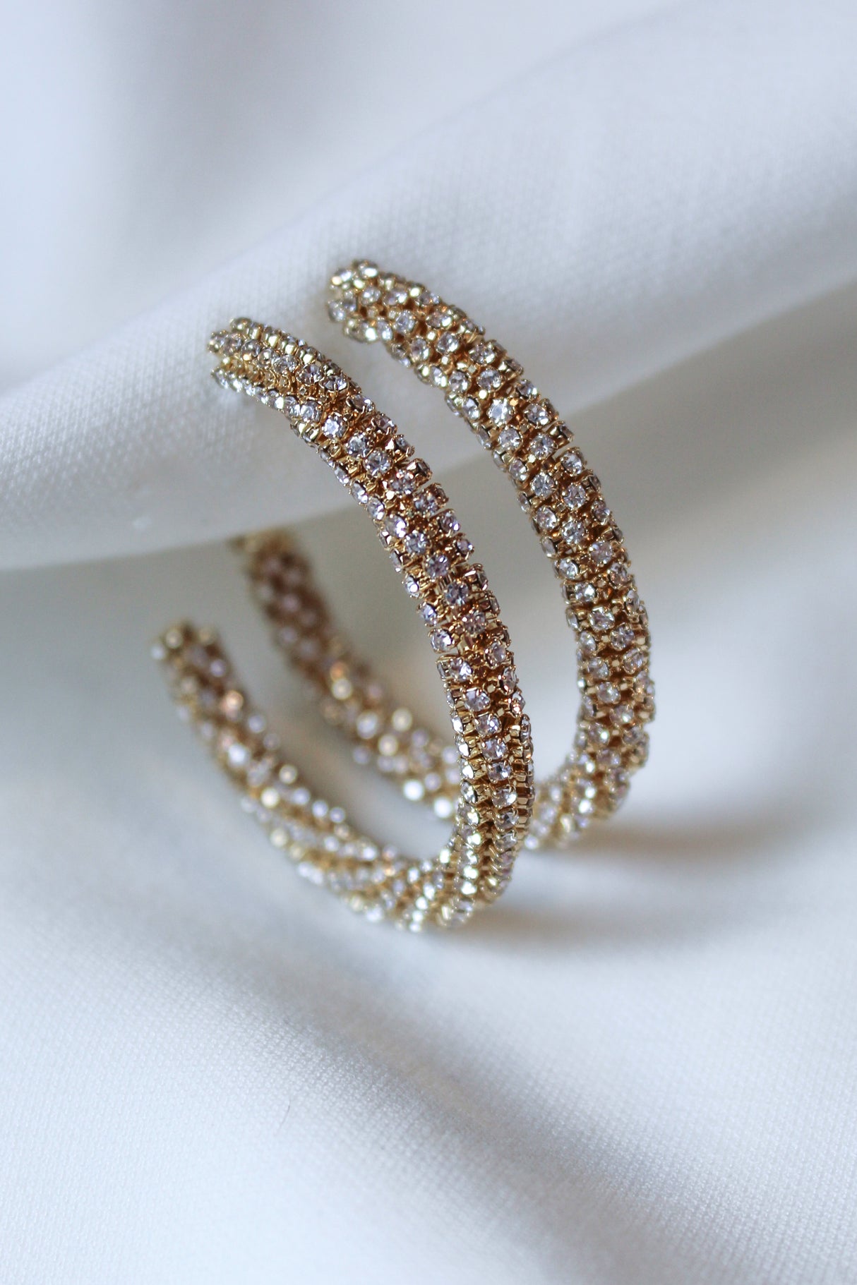 The Riley Hoop by Kinsey Designs features gold-plated, diamond-encrusted double hoops with sparkling cubic zirconia, displayed elegantly on white fabric—an exquisite find at our sample sale.