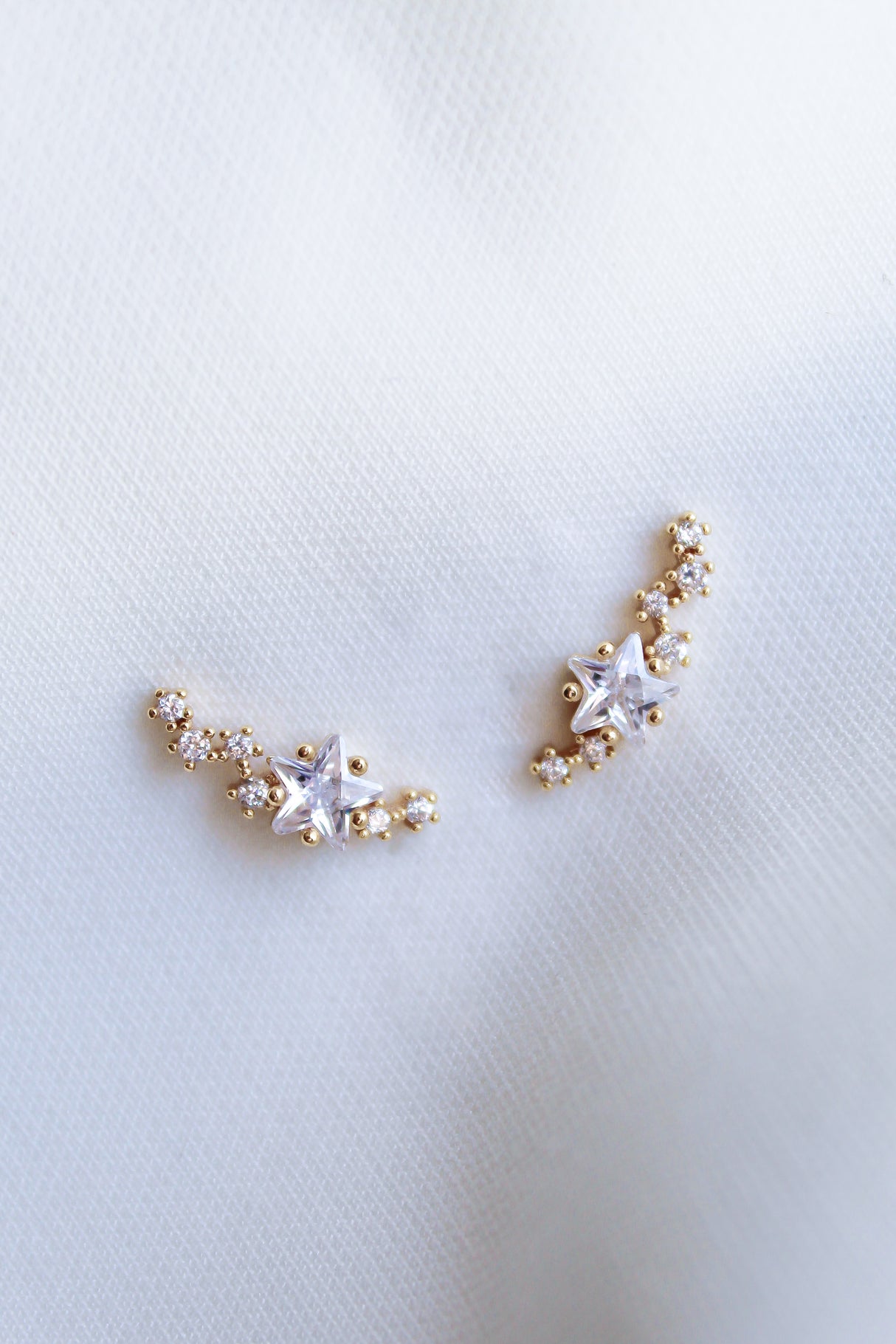 The Kinsey Designs Starlette Climber earrings, featuring star-shaped crystals and sparkling cubic zirconia, are displayed on a soft white fabric background.