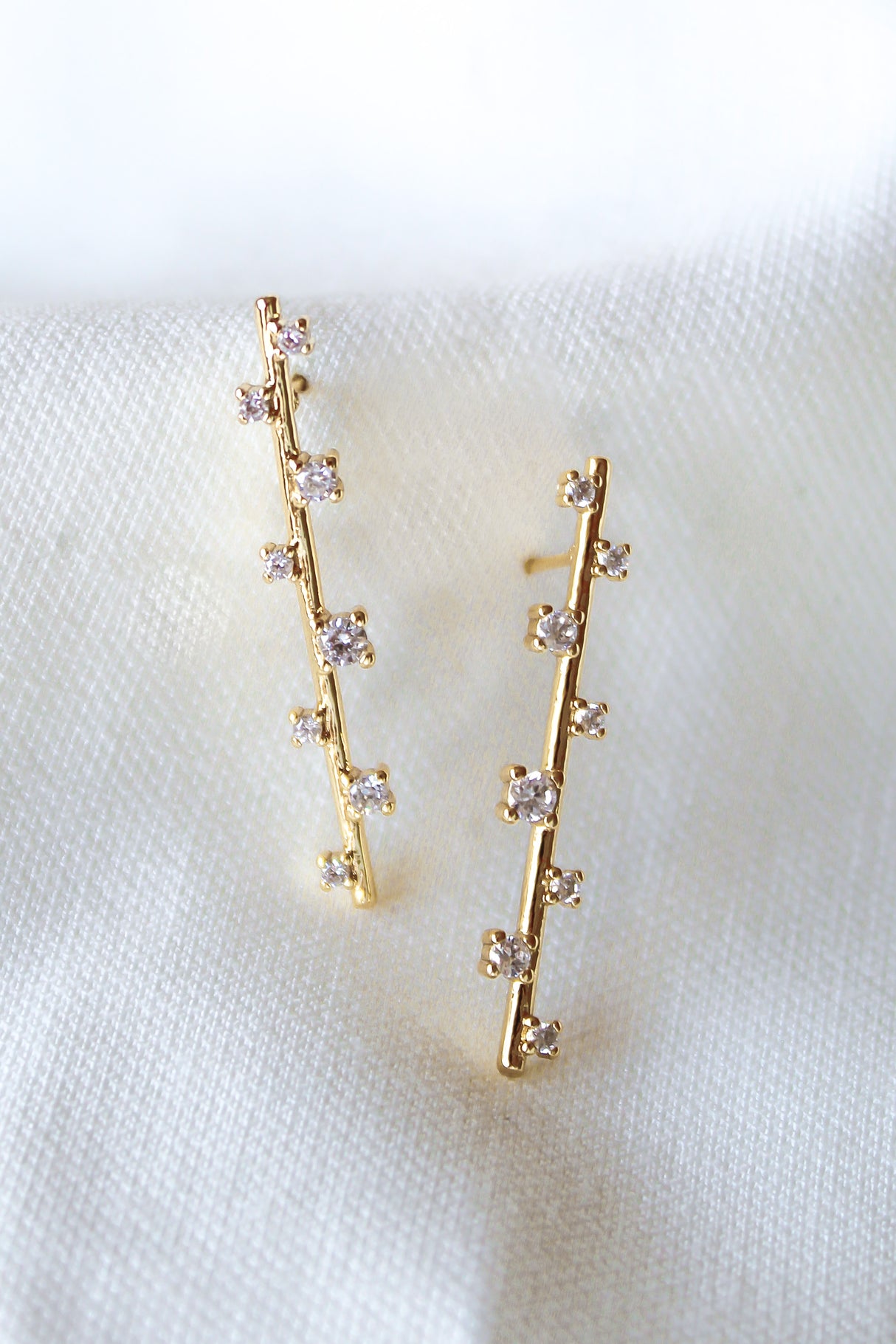 The Walker Earring by Kinsey Designs Archives features 1.5-inch gold-filled drops with vertical rows of small cubic zirconia, shown on a white fabric background. These elegant earrings bring subtle sparkle to any look.