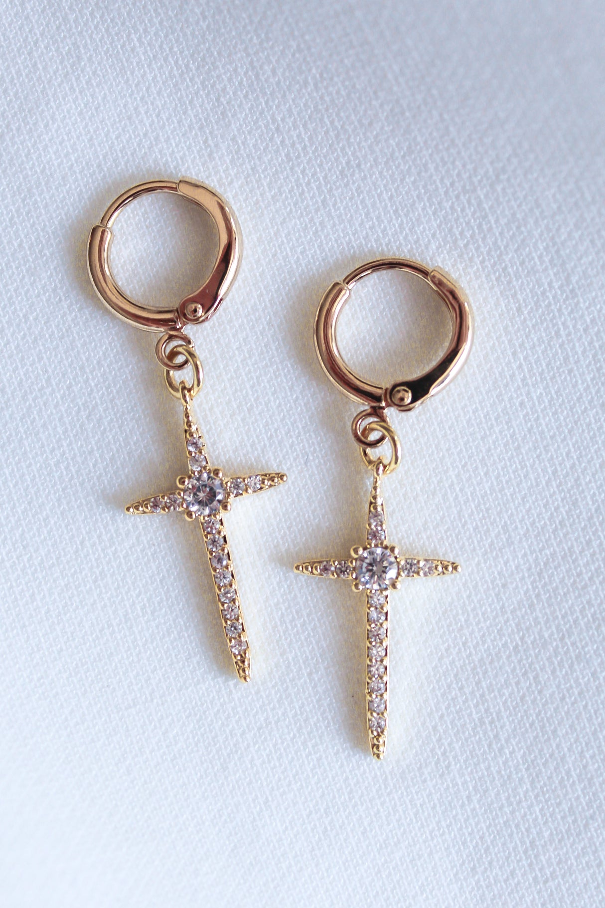 The Zenni Huggie by Kinsey Designs Archives are gold hoop earrings with dangling, jeweled cross pendants featuring sparkling cubic zirconia, displayed on a white textured background.