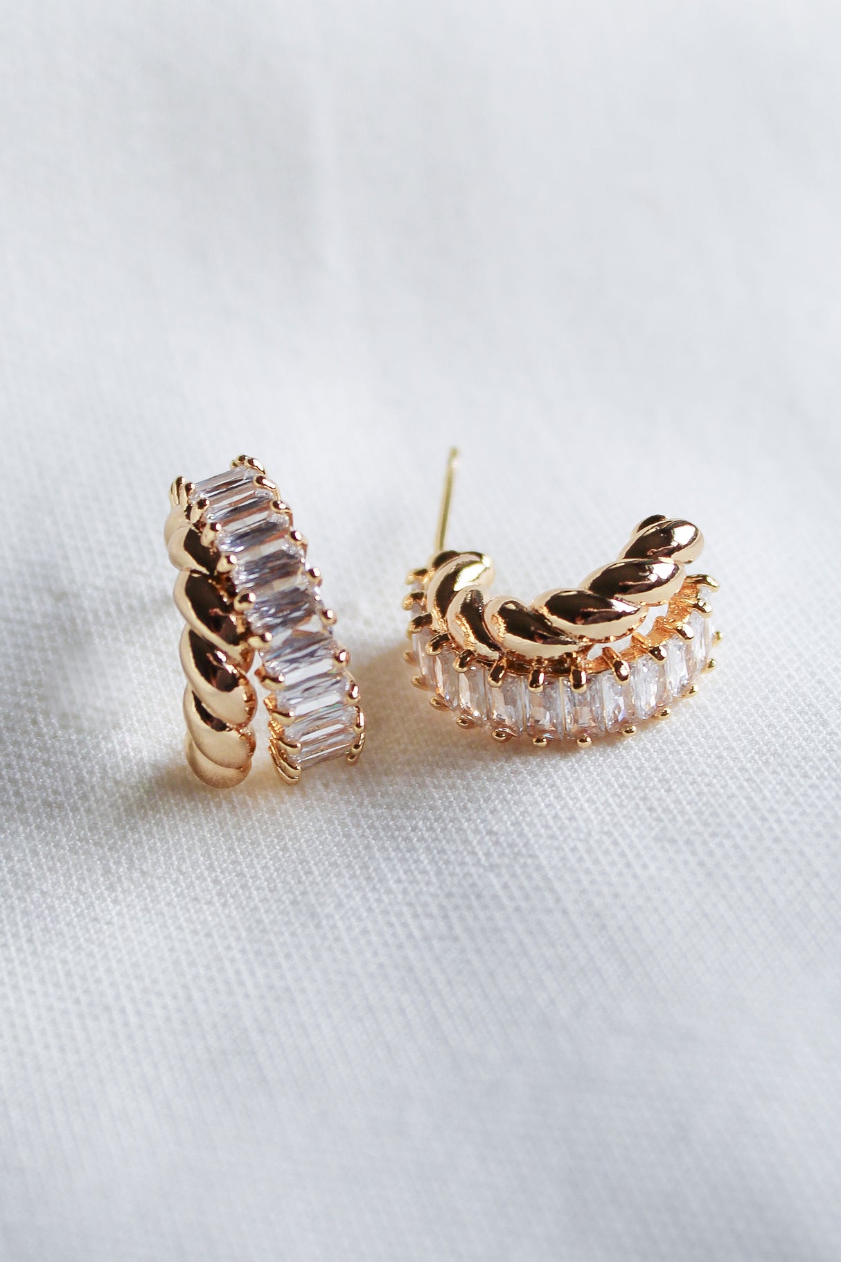 The Zuri Hoop by Kinsey Designs features gold-filled hoops with clear rectangular cubic zirconia in a twisted design, displayed on white fabric.