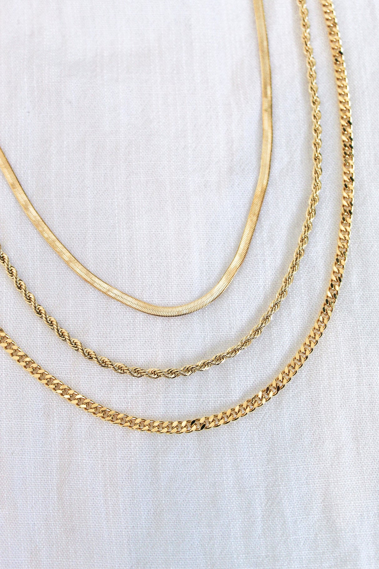 The Amelia Necklace by Kinsey Designs Archives features three gold-plated layered chains in 16" and 18" lengths, each with a unique style, elegantly displayed on a light fabric background.