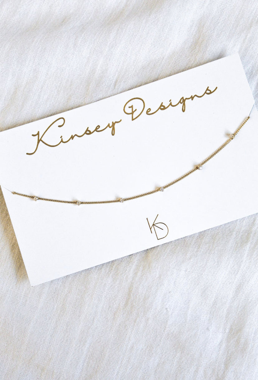 The Aria gold plated necklace by Kinsey Designs Archives, 14 inches long and accented with small gems, is shown on a white Kinsey Designs card against a white fabric background.