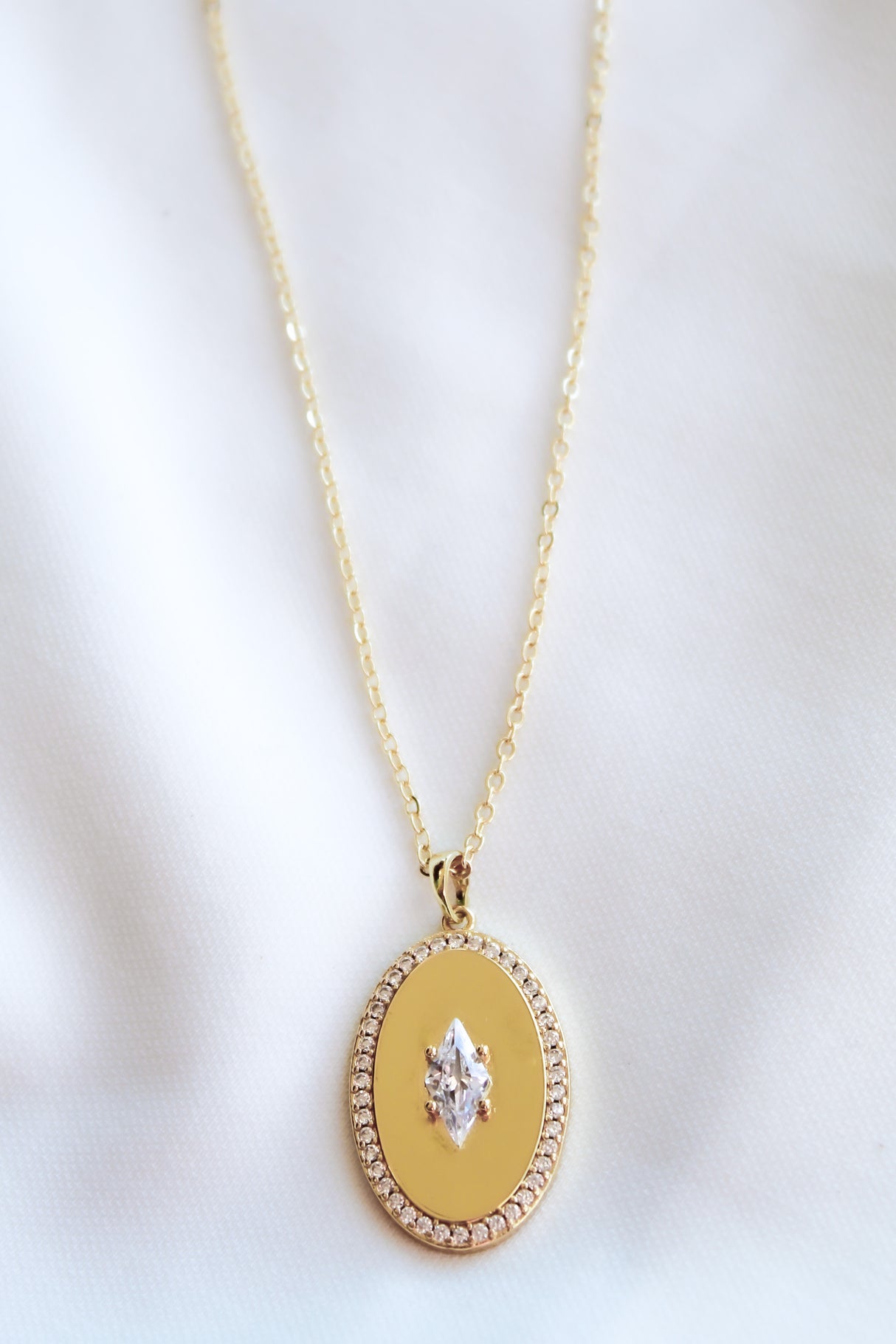 The Fique Necklace by Kinsey Designs is a gold-plated 32-inch chain with an oval cubic zirconia pendant bordered by small diamonds, elegantly displayed on white fabric.