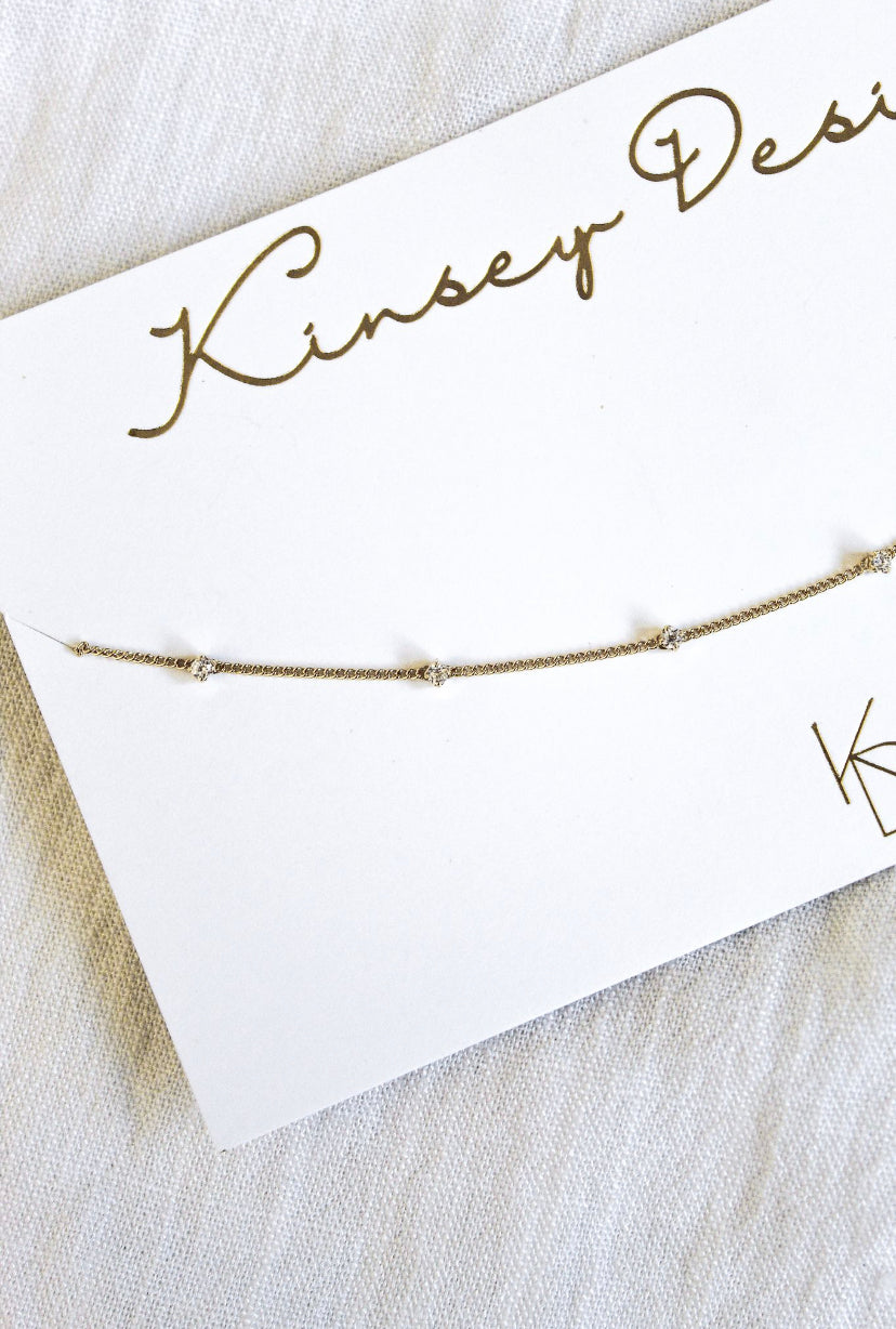 The Aria bracelet by Kinsey Designs Archives features delicate gold plating and small gems. Displayed on a white branded card atop light fabric, it includes a secure lobster clasp for effortless wear.