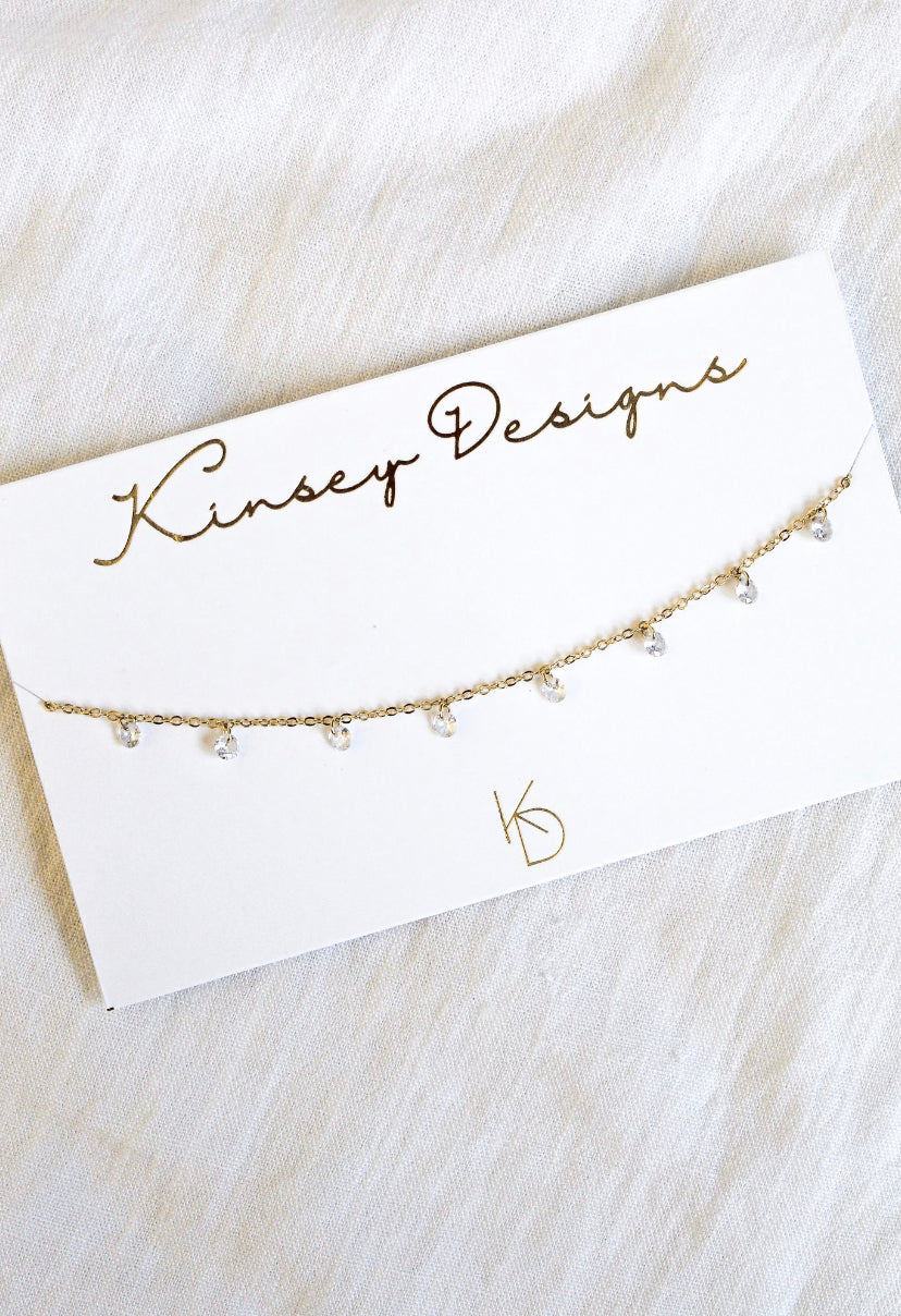 The Clare Choker by Kinsey Designs Archives features a delicate gold-plated 14" chain with small dangling cubic zirconia crystals, presented on a branded card for an elegant, dainty look.
