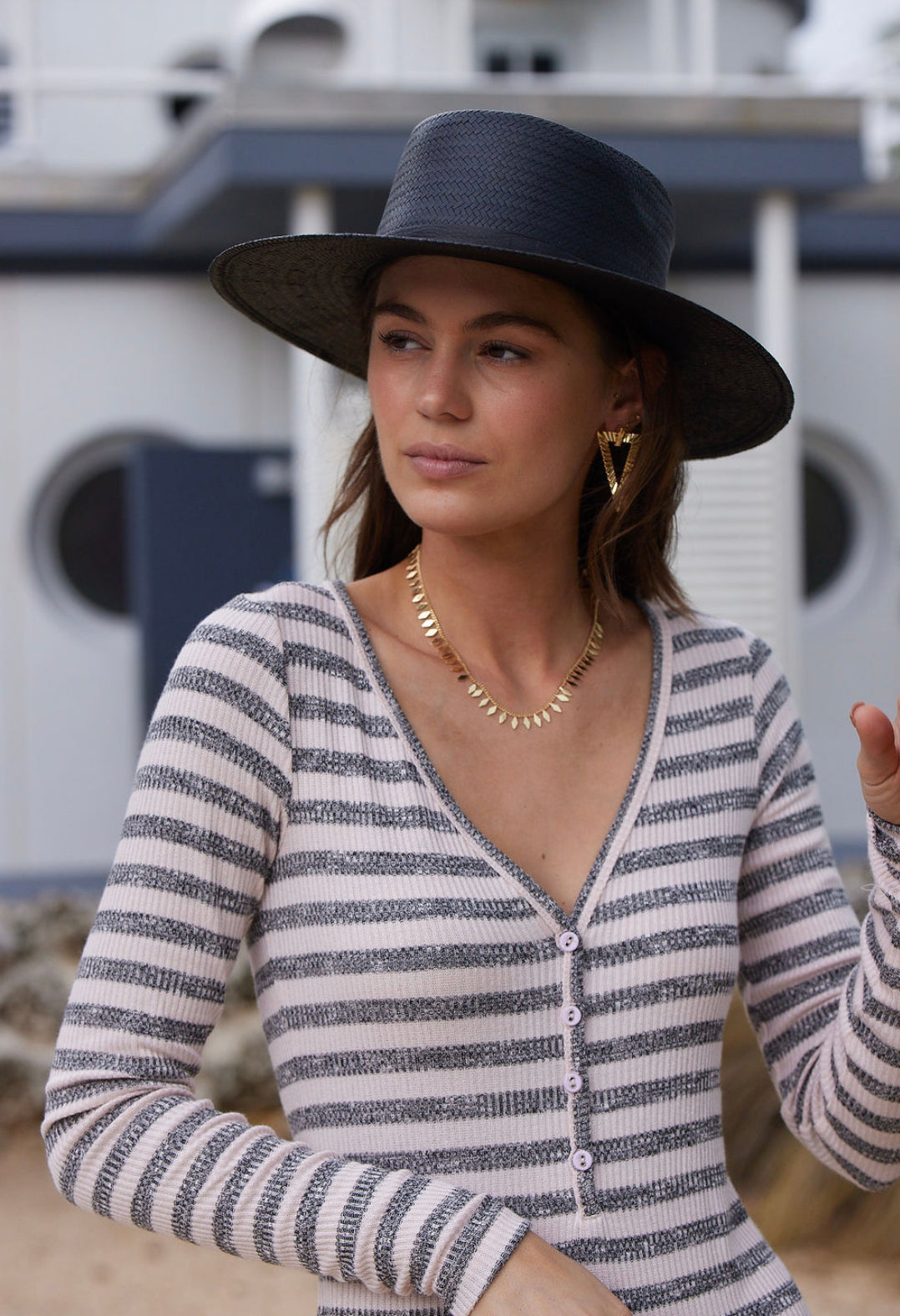 A woman in a striped top and black hat showcases the Fisher Necklace by Kinsey Designs Archives, standing outdoors near a modern building.