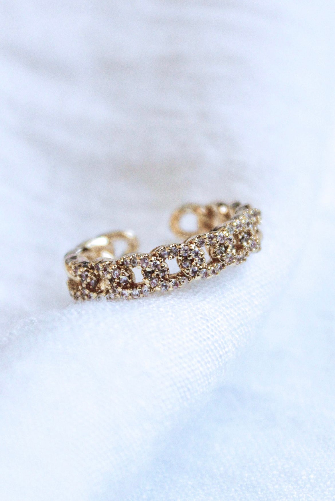 The Ava Lux Ring by Kinsey Designs Archives features a gold-plated, chain-link design adorned with sparkling cubic zirconia, elegantly displayed on white fabric.