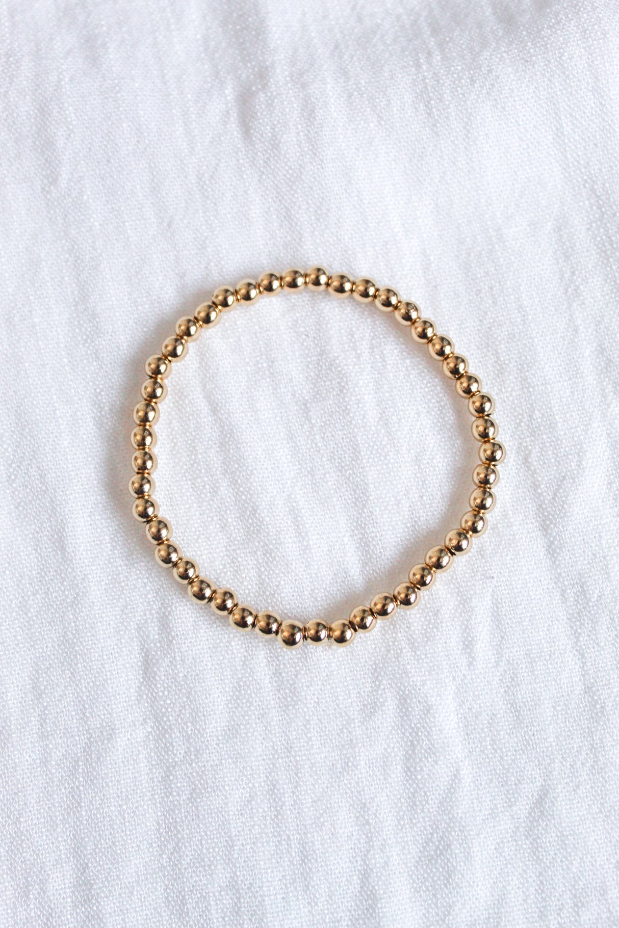 The Kinsey Designs Liam Stacking Bracelet, available in multiple sizes, features gold-filled beads and an adjustable stretch fit, shown arranged in a circle on a white fabric background.