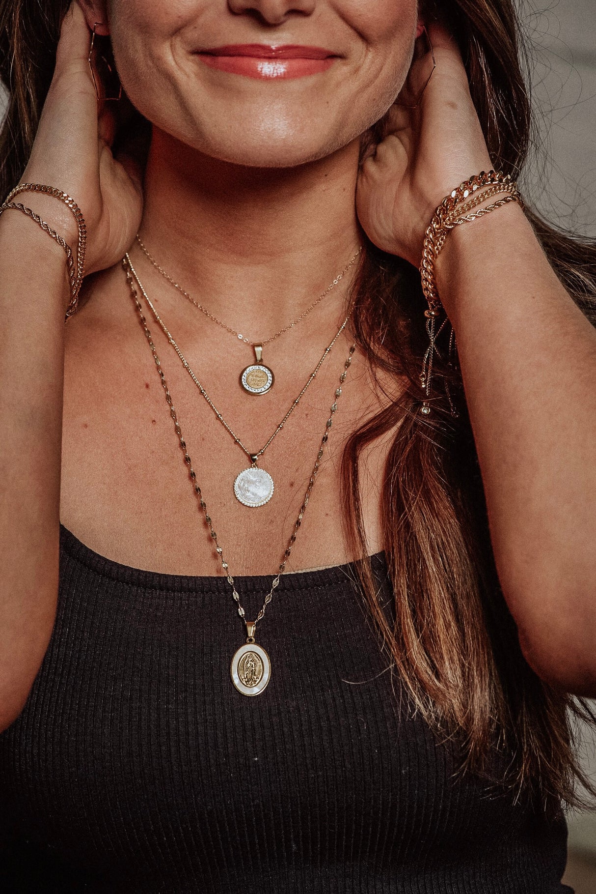A woman smiles with her hands near her face, wearing the Kinsey Designs Archives Alivia layered 14k gold plated necklaces with a mother of pearl charm, styled with a black top.