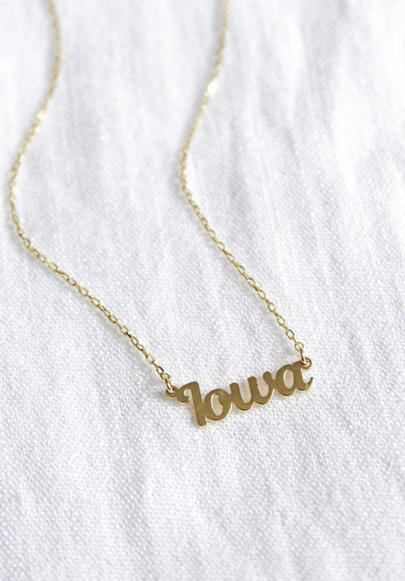 The State of Mind Necklace by Kinsey Designs Archives features a gold filled “Iowa” cursive pendant, shown on a white textured fabric background.