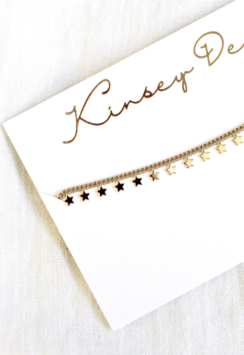 The Starry Choker by Kinsey Designs Archives features a gold plated chain with delicate star charms, displayed on a white "Kinsey D" card—a stylish, elegant addition to your jewelry collection.