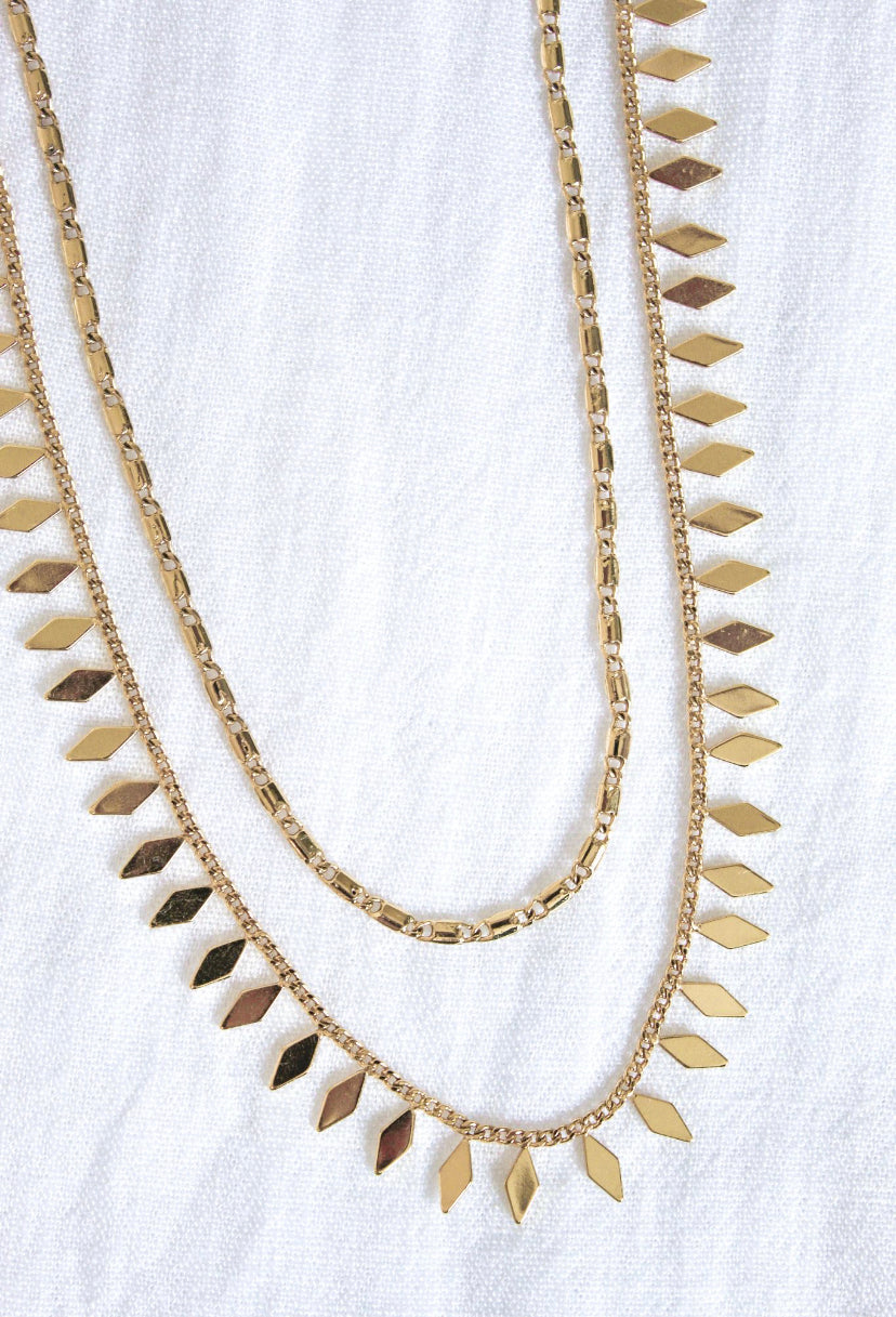 The Fisher Necklace by Kinsey Designs Archives features two layered 14k gold plated chains with small diamond-shaped pendants, displayed on a white textured fabric background.