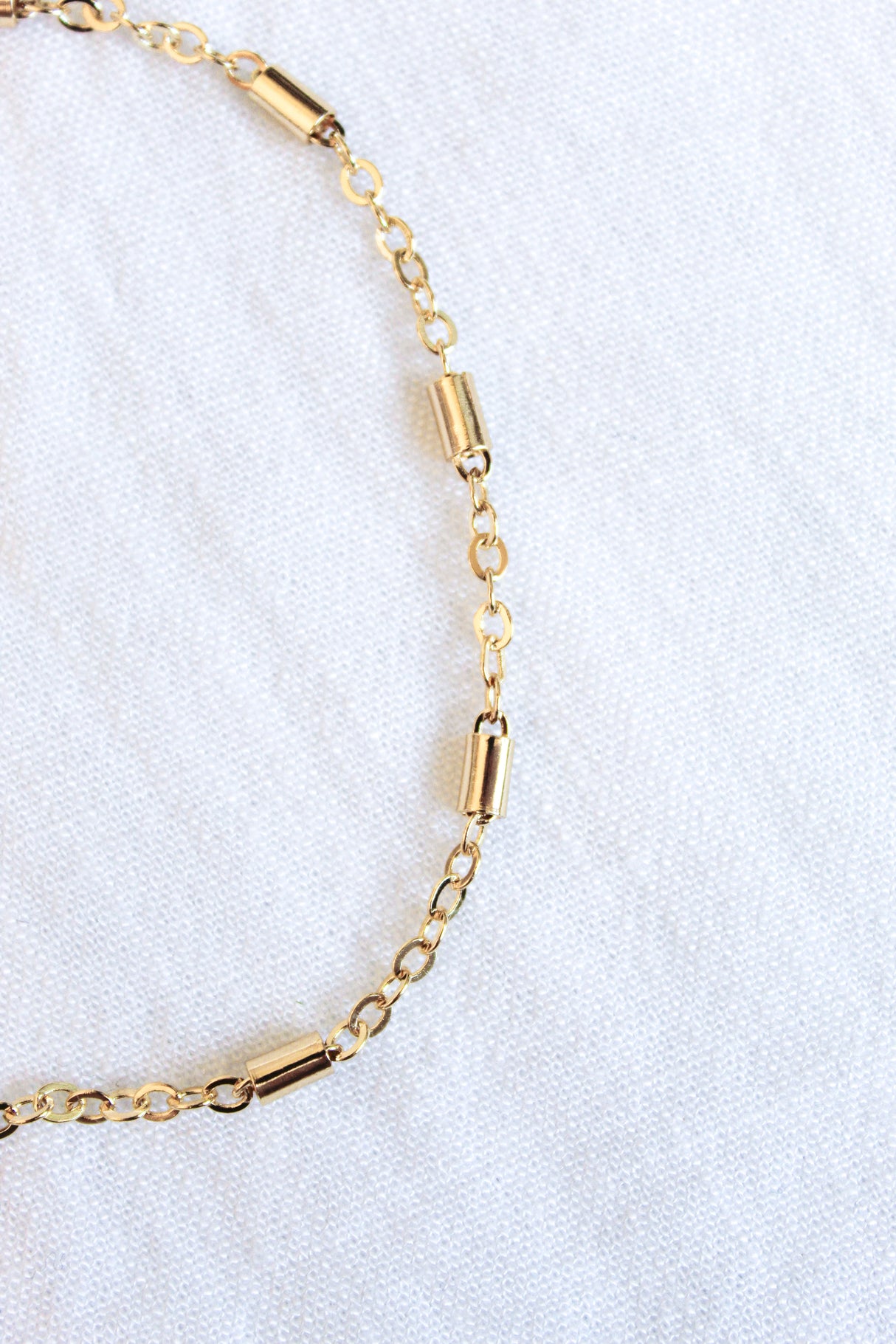 The gold chain bracelet with cylindrical beads rests on a white textured fabric, paired with the Kinsey Designs Archives Azure Anklet - Gold Filled for added elegance.