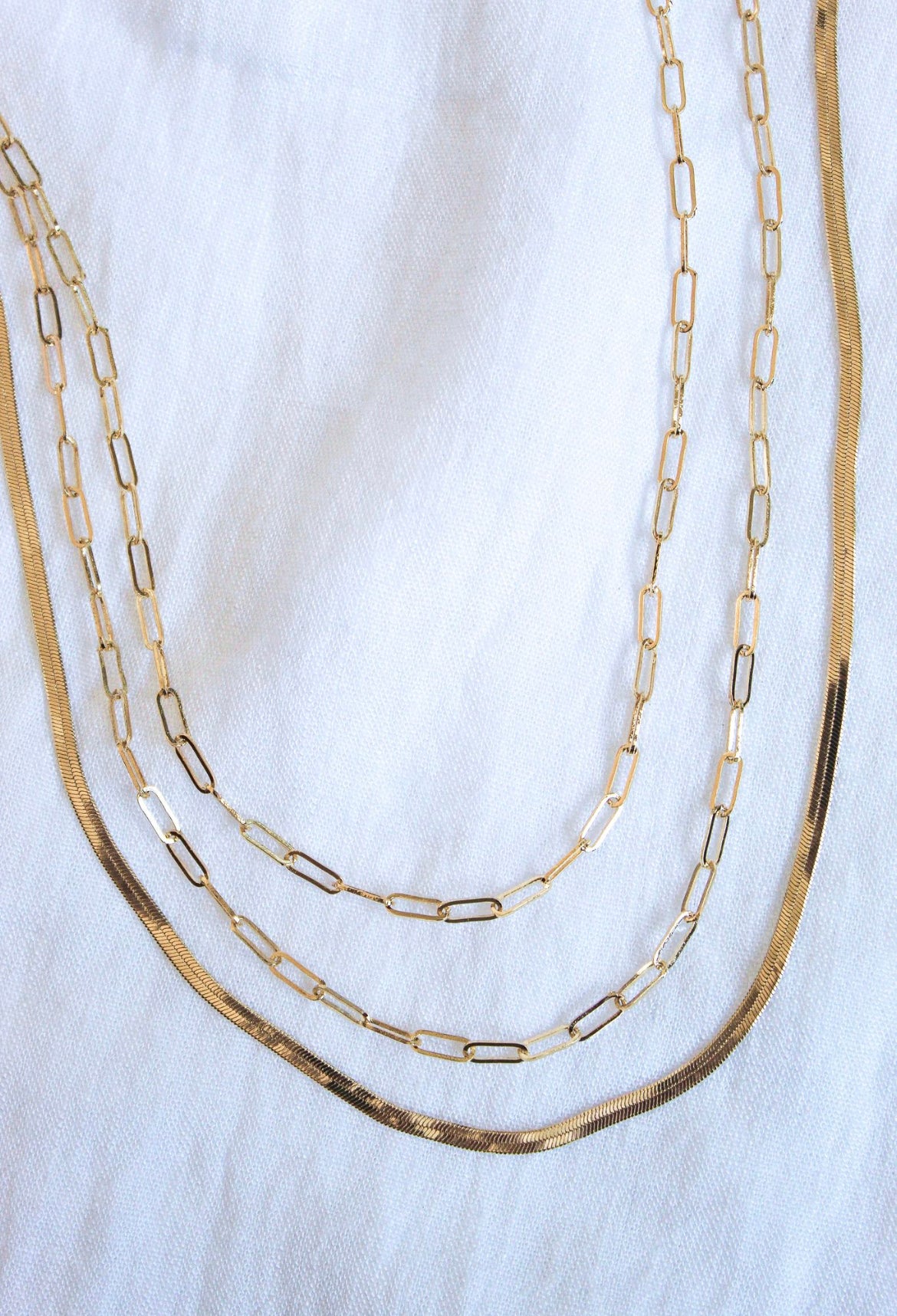 The Wren Chain Layer by Kinsey Designs Archives, a three-layered 16k gold-plated brass necklace with rectangular links, is elegantly displayed on a white fabric background.