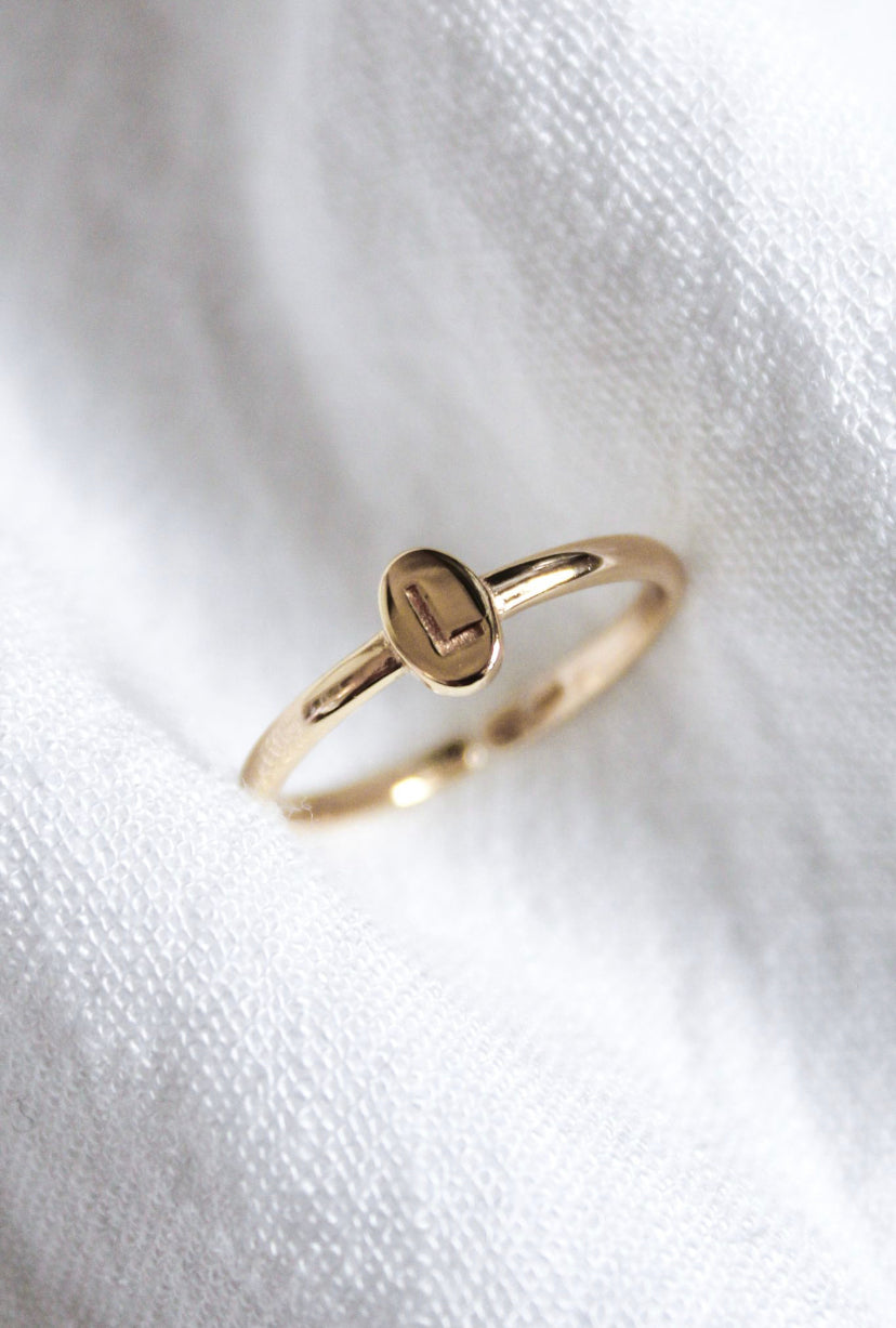 The Kinsey Initial Ring by Kinsey Designs, engraved with the letter "L," rests on a soft white fabric background, making it a stunning piece of personalized jewelry.