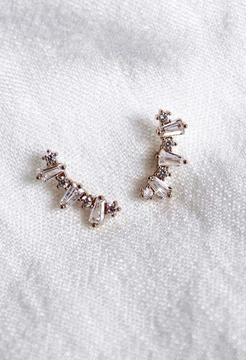 The Irace Post earrings by Kinsey Designs Archives feature small cubic zirconia stones in a curved line, displayed on white textured fabric.