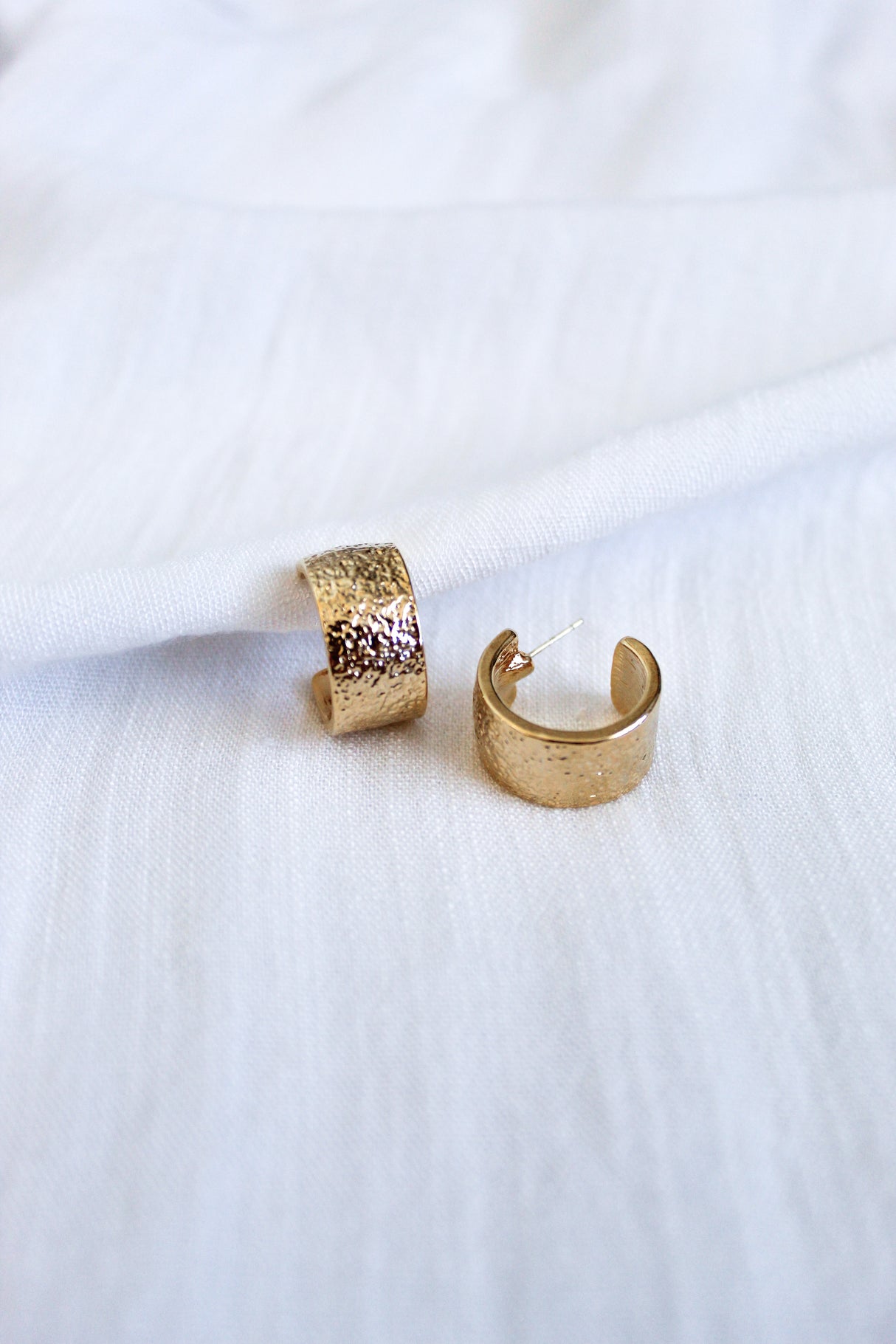 Two Fawn Hoop gold-plated brass earrings by Kinsey Designs Archives, featuring a textured finish, rest elegantly on a white fabric background.