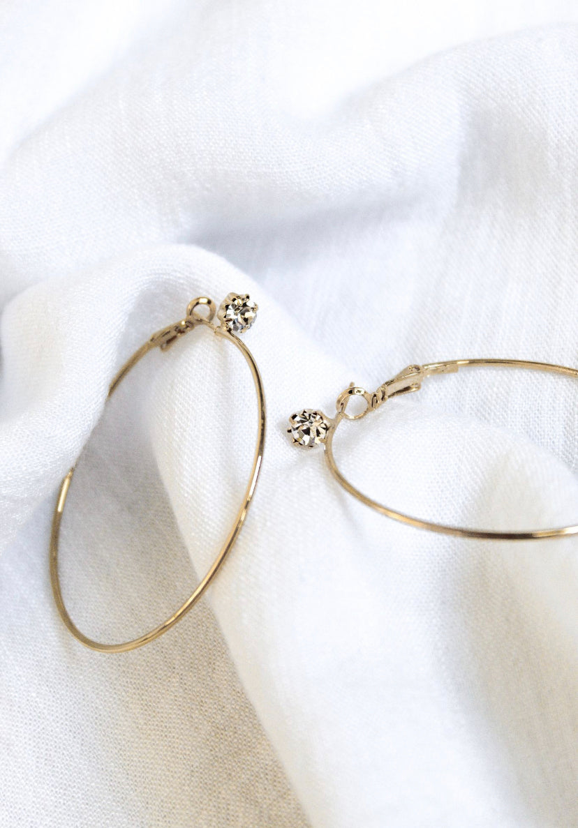 The Ashton gold plated hoop earrings by Kinsey Designs Archives, 2” wide and accented with small cubic zirconia, are displayed on a soft white fabric background.