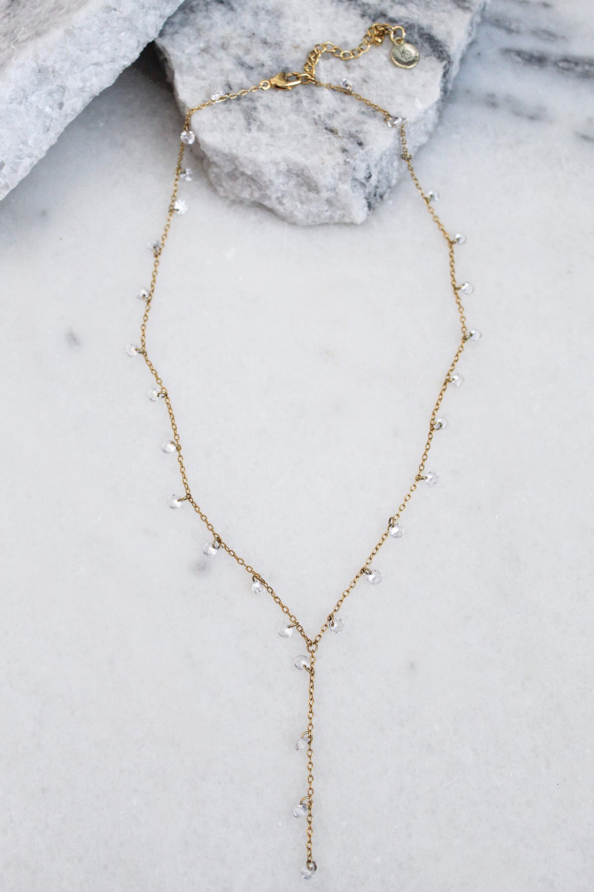 The Clare Lariat by Kinsey Designs Archives is an 18k gold plated necklace with small clear beads, CZ charms, a drop pendant, and an adjustable extender. Shown on marble, it adds elegance to any look.