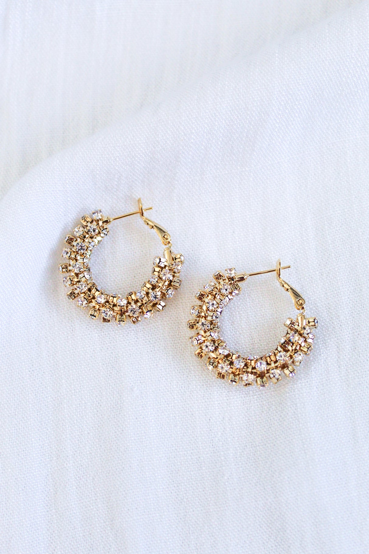 Kinsey Designs' Harkness Hoops, featuring sparkling cubic zirconia, are showcased against a white textured fabric backdrop.
