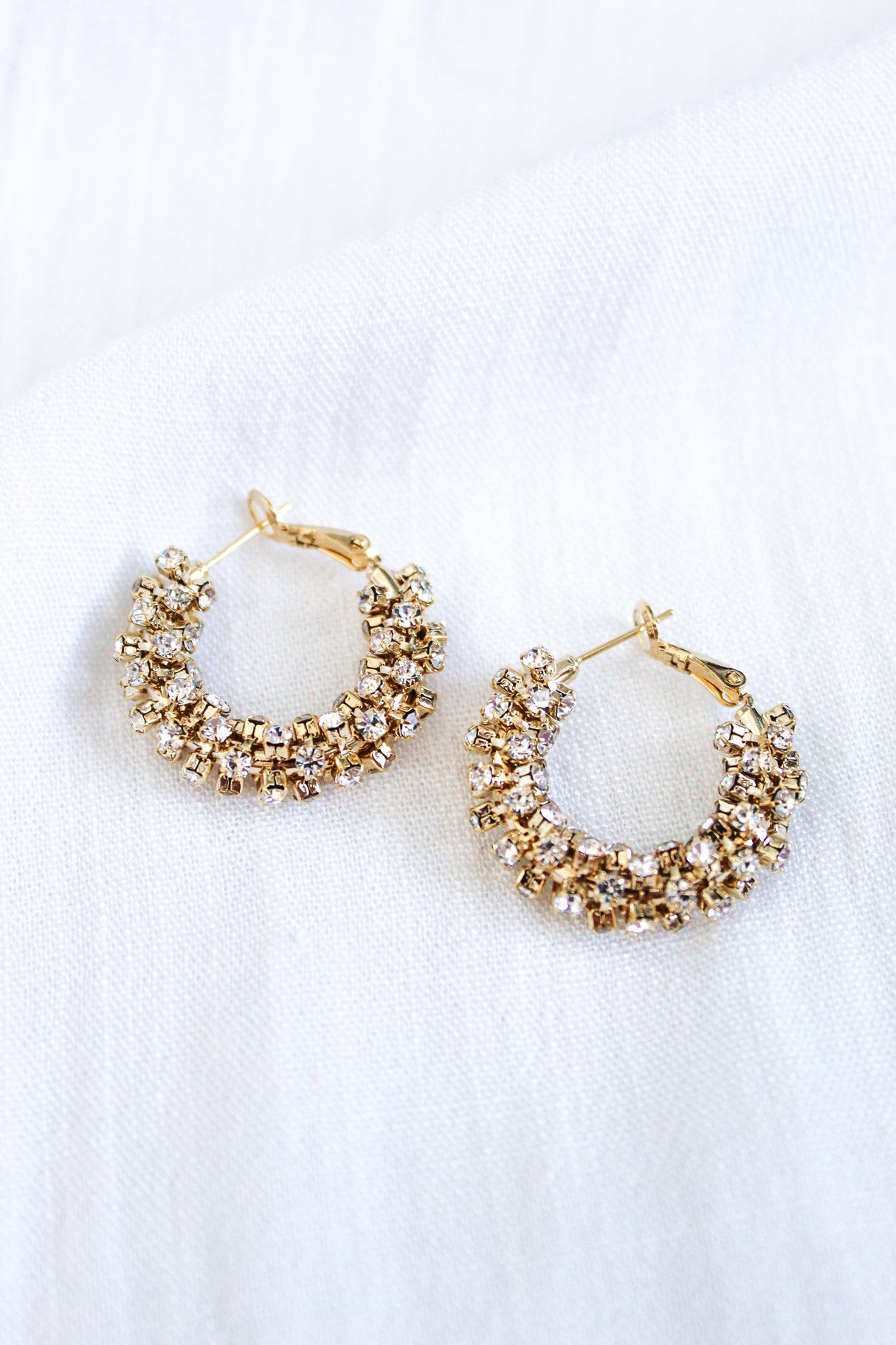 The Harkness Hoops by Kinsey Designs feature gold crystal hoops adorned with clusters of sparkling cubic zirconia, shown against a white fabric background.