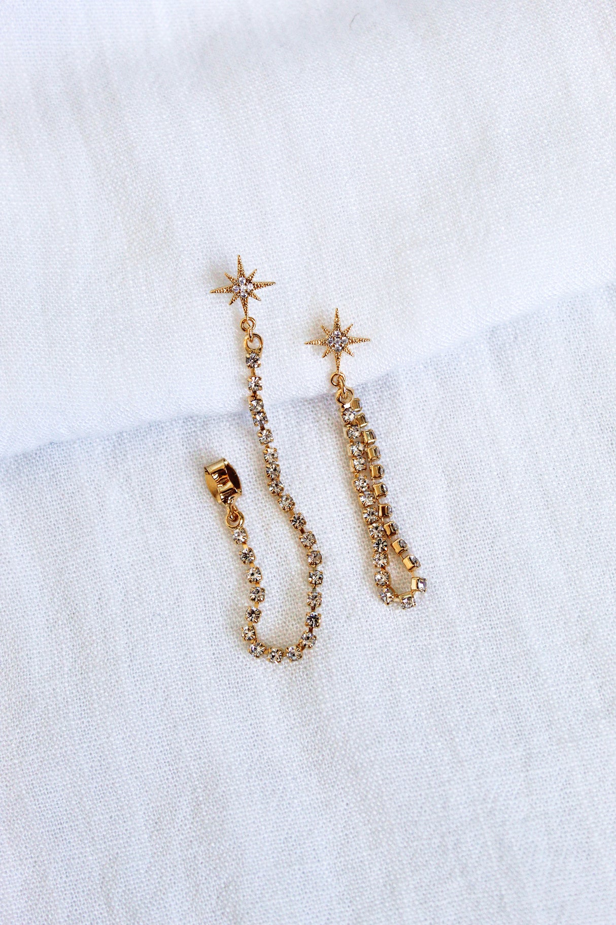 The Reese Drop Earrings by Kinsey Designs Archives are gold plated, star-shaped with crystal chains, shown on a white textured fabric background.