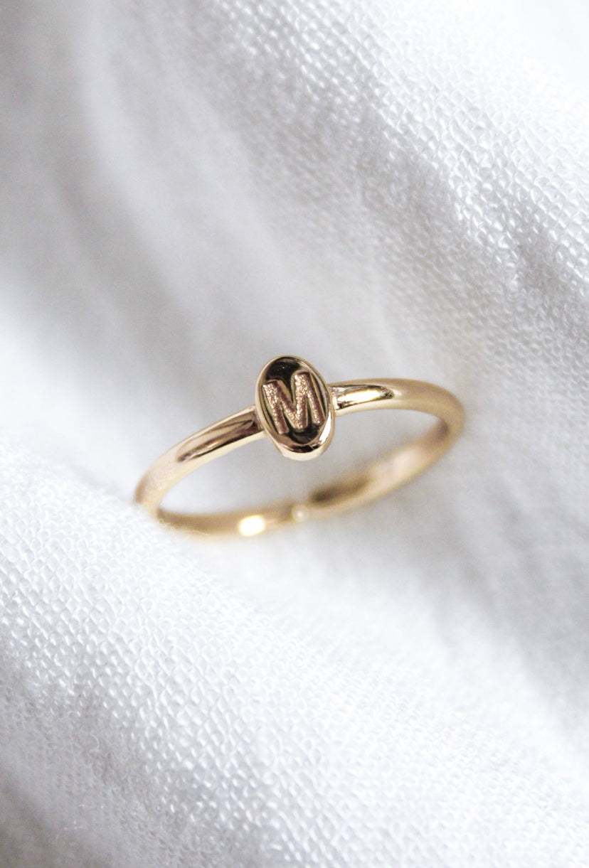 The Kinsey Initial Ring by Kinsey Designs features an oval gold face engraved with the letter "M," displayed on soft white fabric—a timeless, personalized jewelry piece.