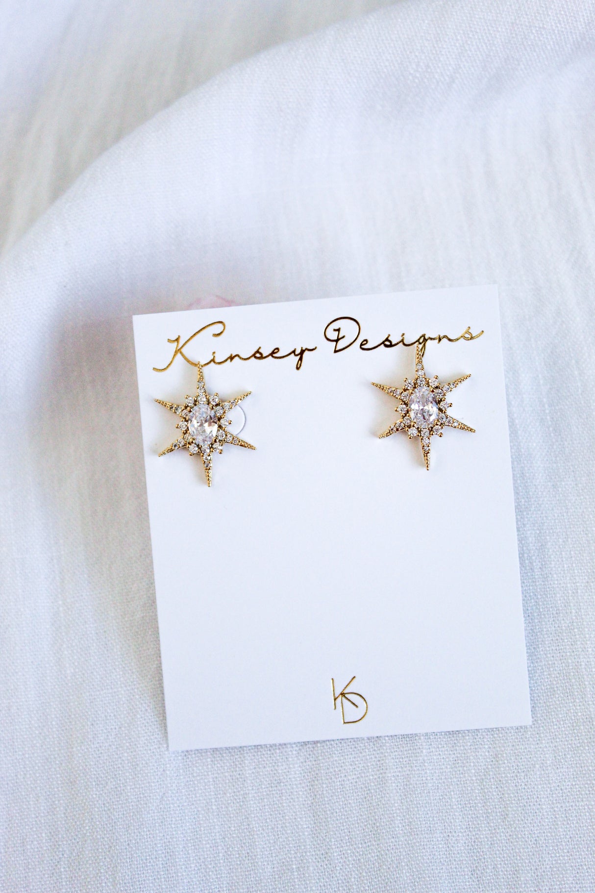 The Faye Stud gold star-shaped earrings by Kinsey Designs feature sparkling cubic zirconia on a branded card, displayed against light fabric. Add shine to your style—shop the sample sale now!.