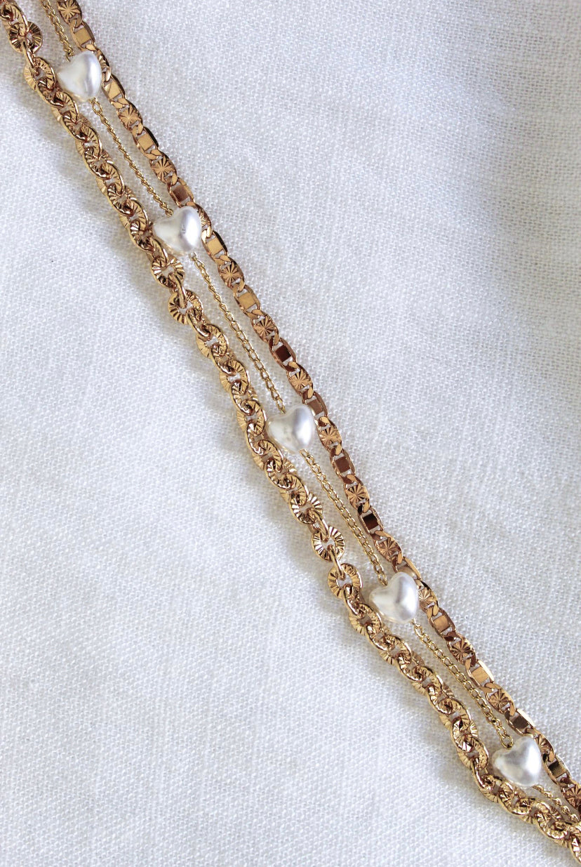 The Beyah Layer Bracelet by Kinsey Designs Archives features gold-plated brass chains with small pearls, displayed diagonally on a white textured fabric background.