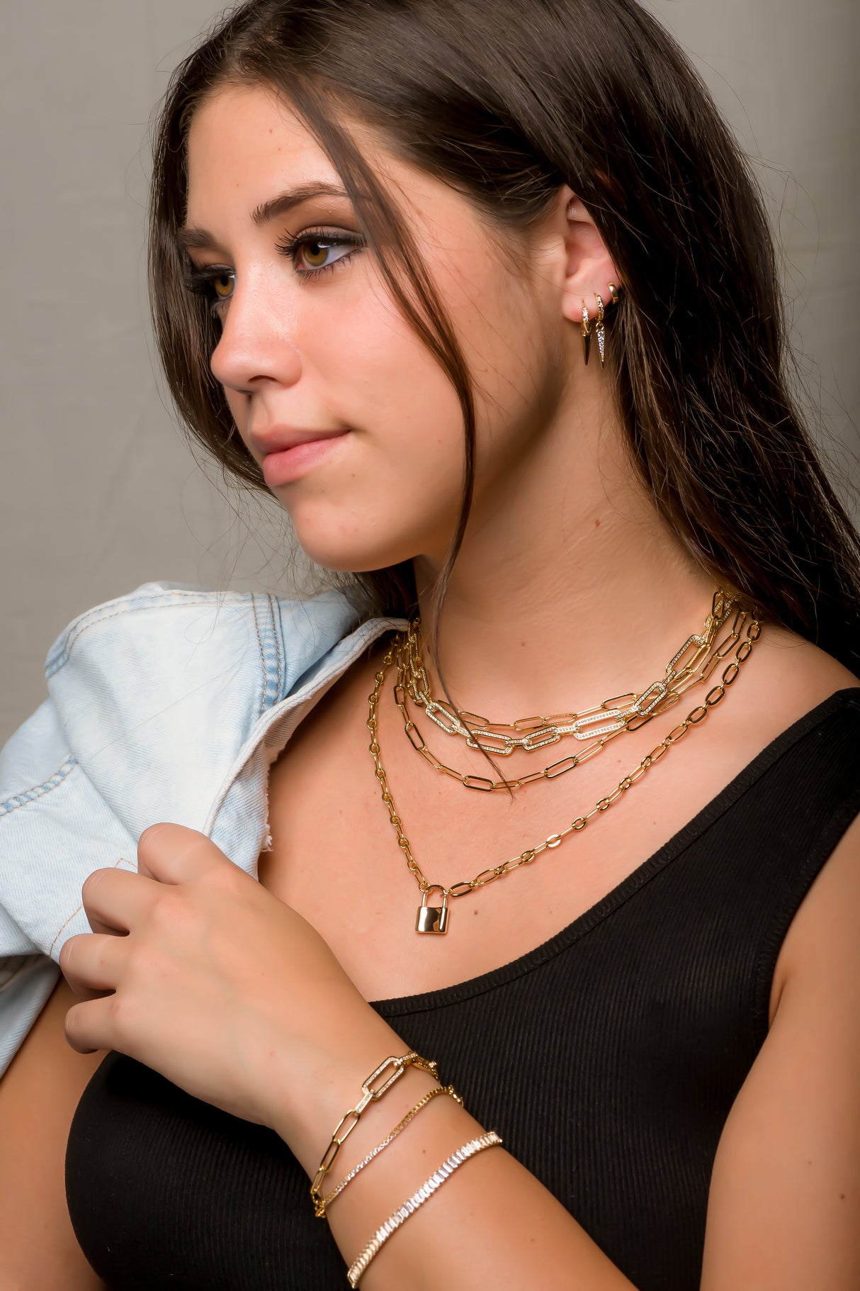 Woman with long brown hair wearing gold jewelry, including the Maeve Necklace by Kinsey Designs, a black top, and a light denim jacket draped over her shoulder.
