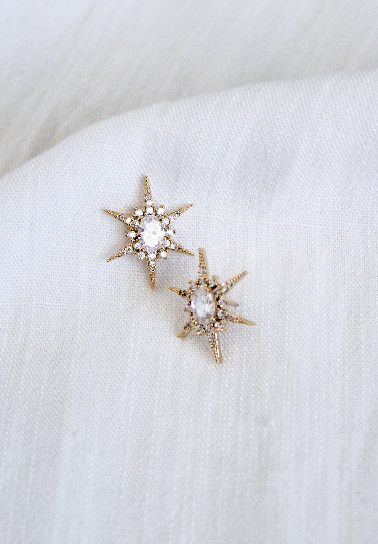 The Faye Stud earrings by Kinsey Designs feature gold-filled, star-shaped studs with clear cubic zirconia centers, displayed on a white textured fabric background.