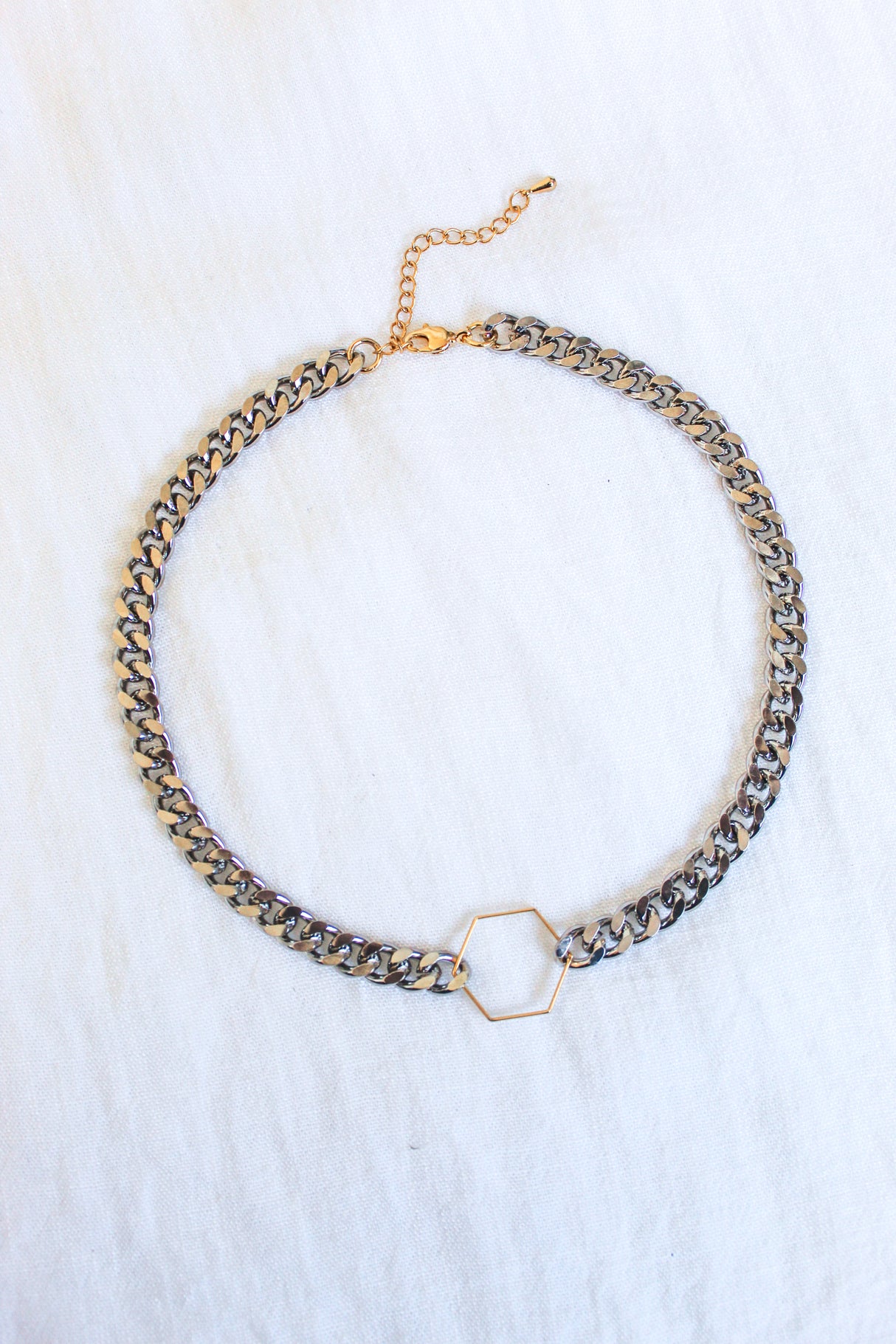 The Fraser Necklace by Kinsey Designs Archives features a 17-inch silver chain with a gold hexagon pendant, crafted from rhodium-plated brass and finished with an adjustable clasp and extender, displayed on white fabric.