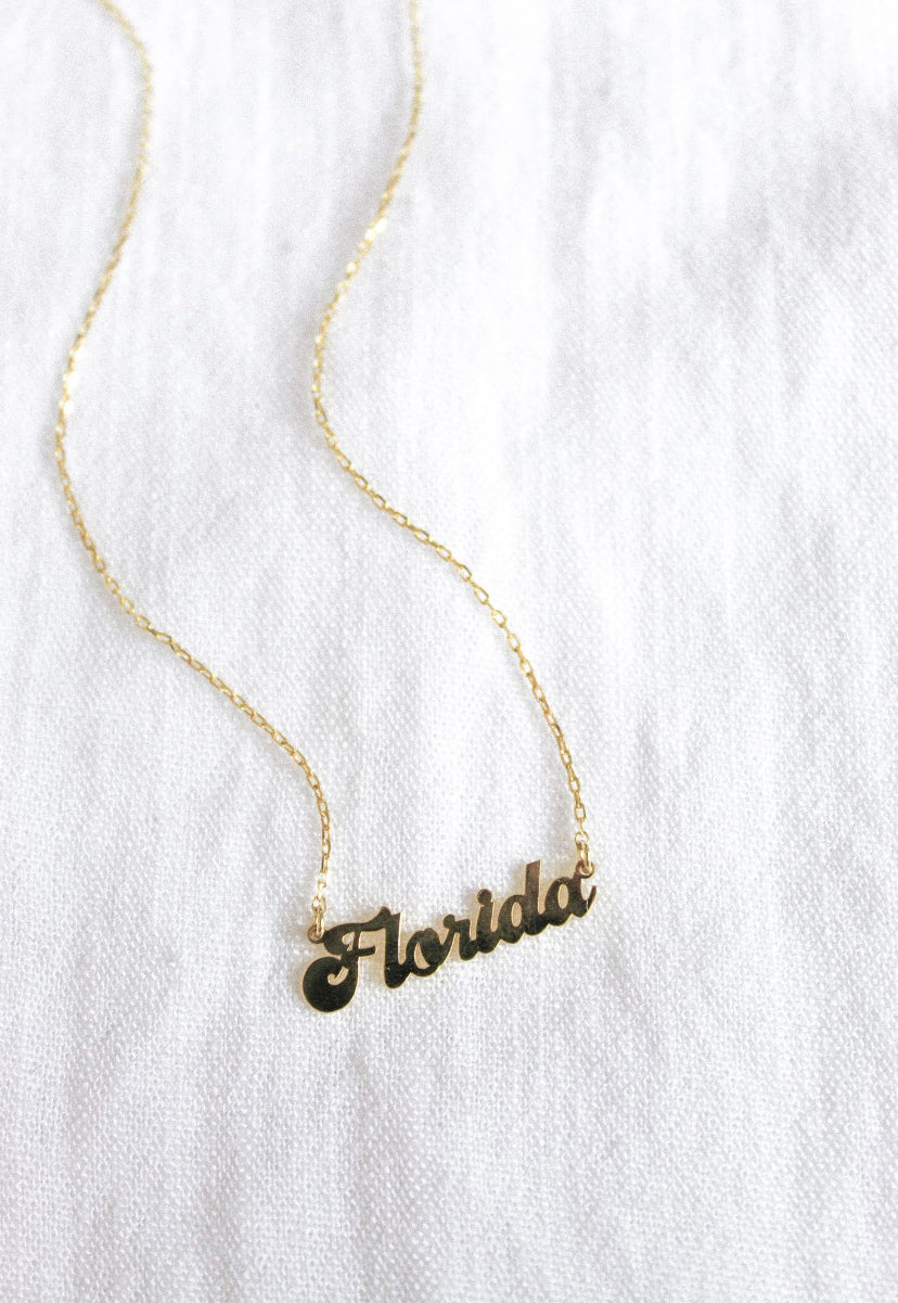 The Kinsey Designs Archives State of Mind Necklace features a gold filled "Florida" cursive pendant, shown on a white fabric background.