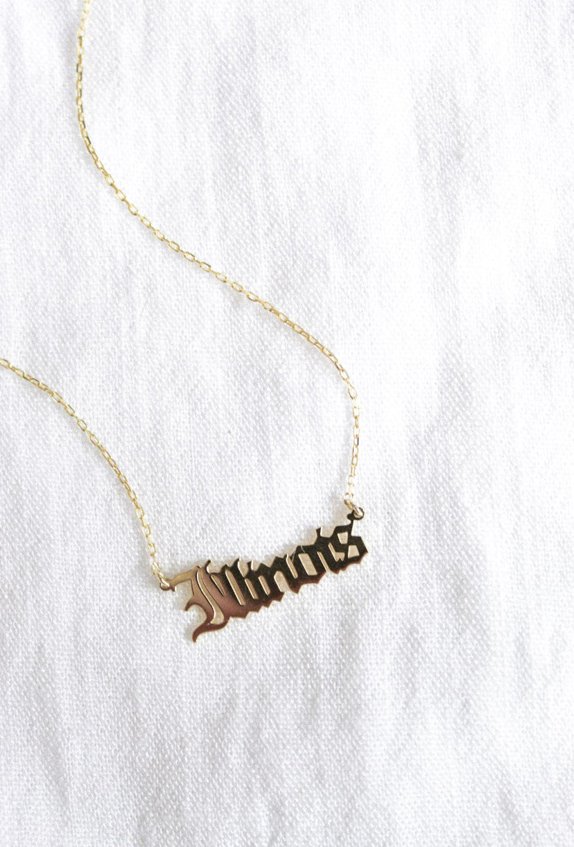 The Kinsey Designs Archives State of Mind Necklace features a gold filled pendant hand stamped with "Illinois," shown on a white textured fabric background.