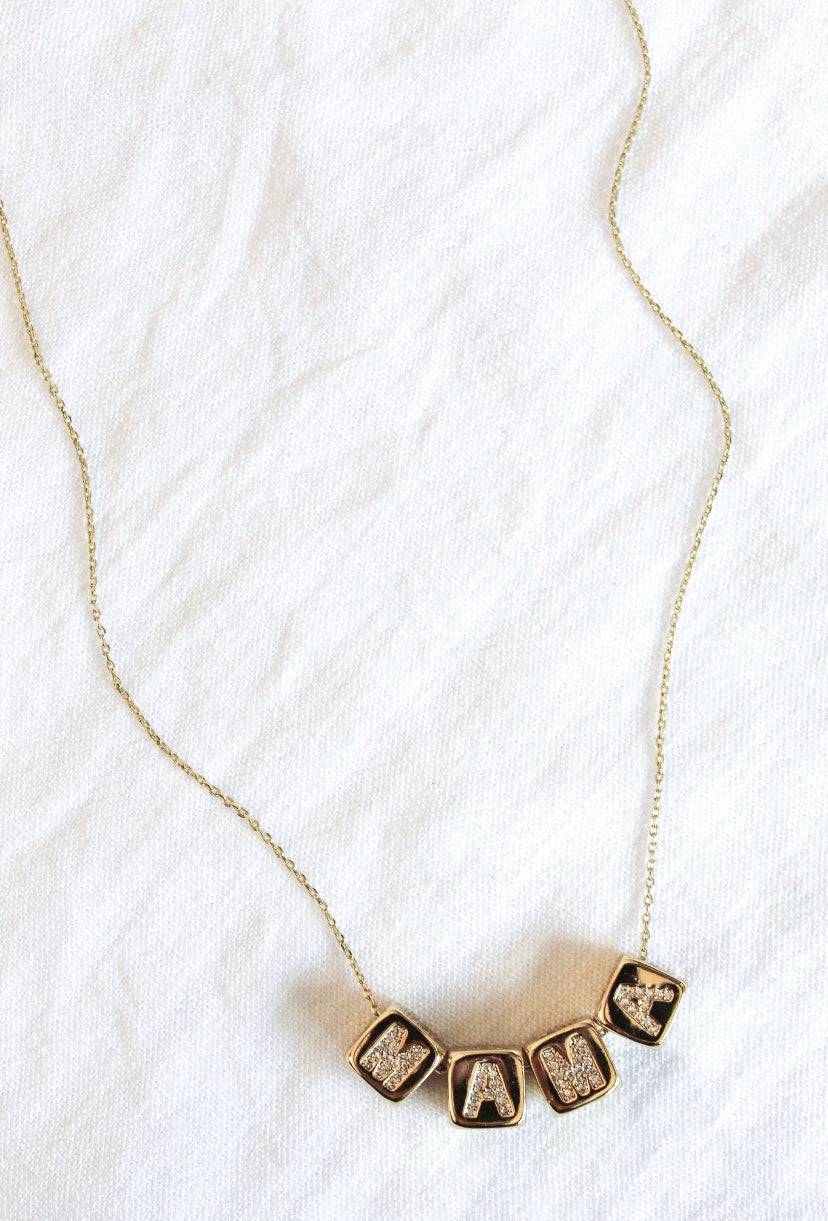 The Kinsey Designs MAMA Block Necklace features gold lettered blocks spelling "MAMA," displayed on a white textured fabric background—a stylish mama necklace and perfect Mother’s Day gift.