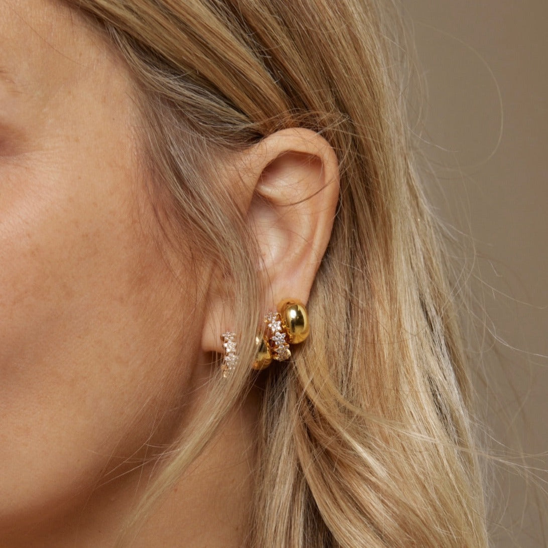 Close-up of a woman's ear with blonde hair, wearing Kinsey Designs Archives' Cloud Huggie gold and diamond stud earring, paired with a delicate gold-plated ring adorned with sparkling cubic zirconia.