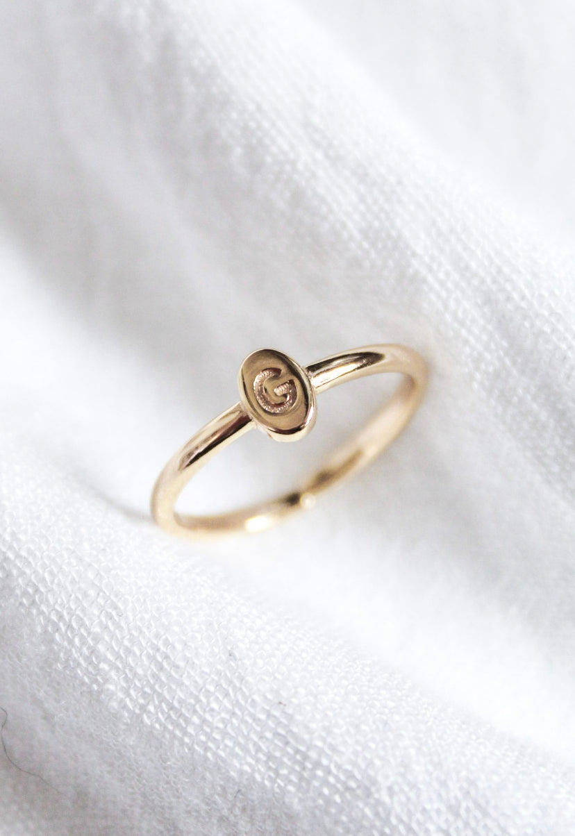 The Kinsey Designs Collection Kinsey Initial Ring Letter G features a gold band with a small oval top engraved with the letter "G," shown on white fabric.