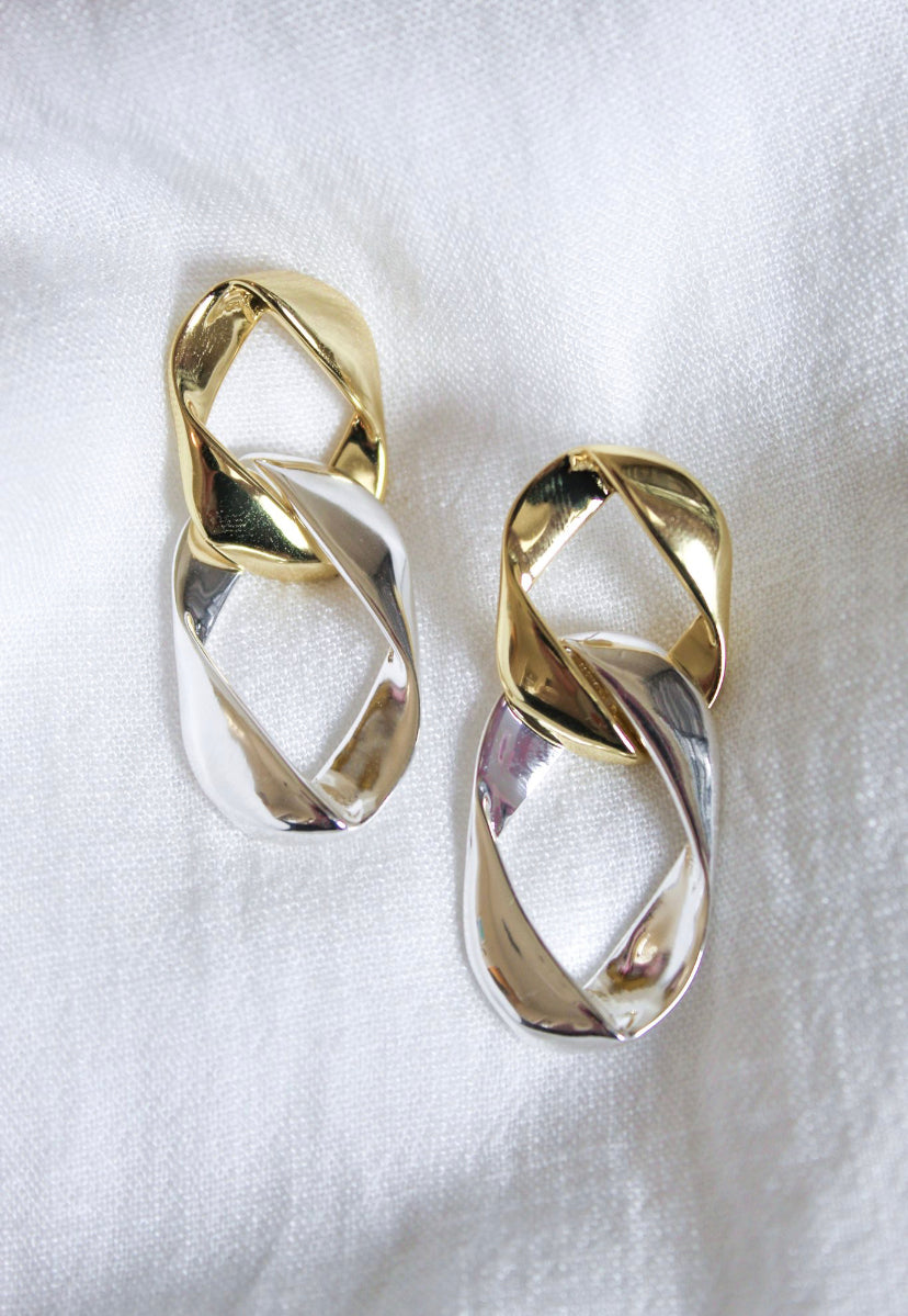 The Porter Earring by Kinsey Designs Archives features two interlocked metallic earrings—one 14k gold plated and one silver over brass, each 2 inches long—displayed on a white fabric background.