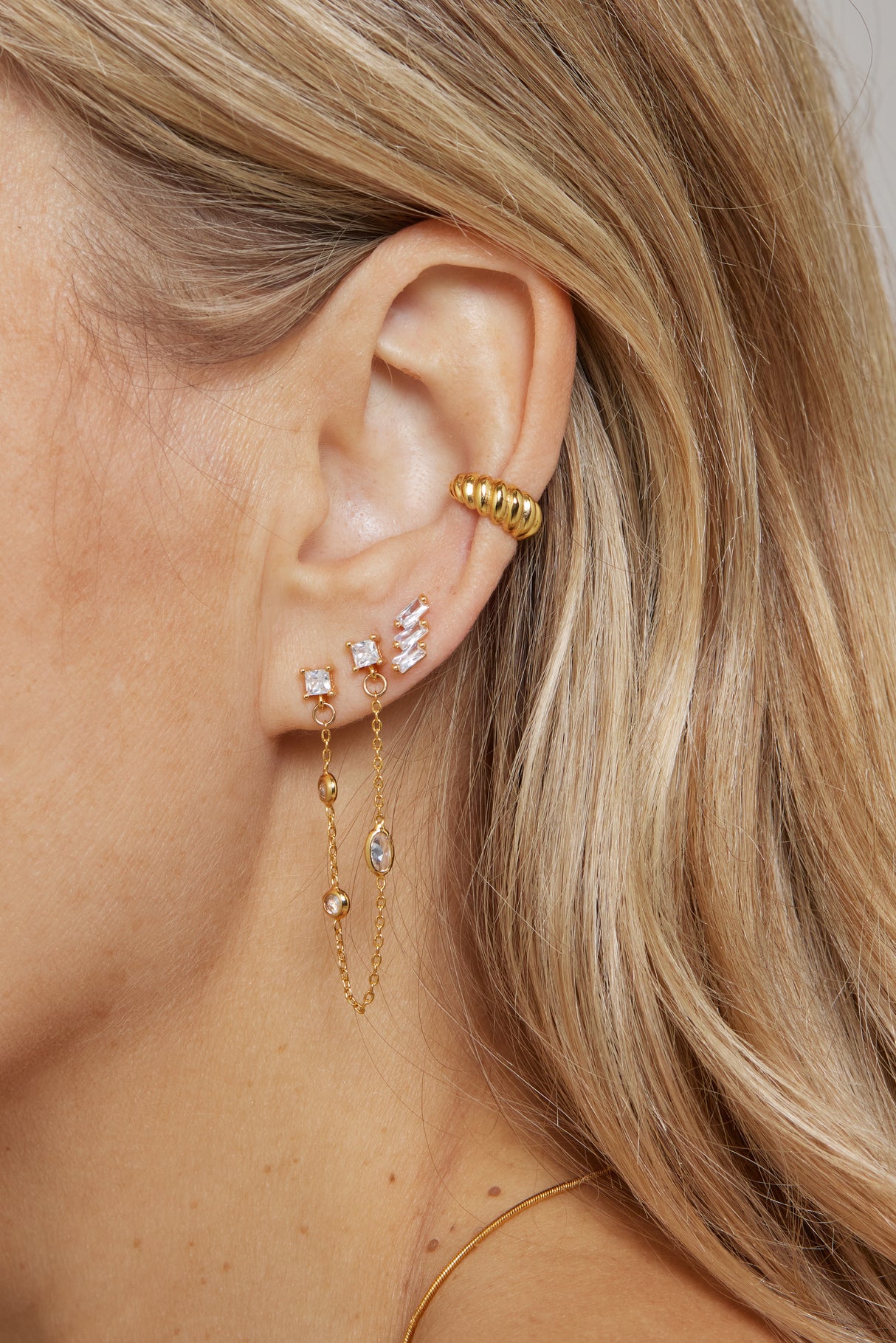 Close-up of a woman's ear adorned with multiple gold-plated and diamond earrings, featuring the Metz Cuff by Kinsey Designs Archives, along with a chain earring and an ear cuff.