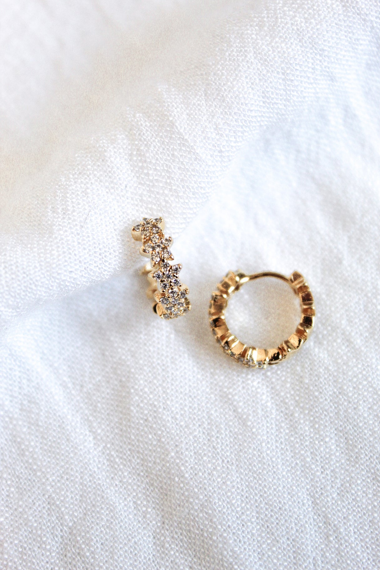 The Cloud Huggie earrings by Kinsey Designs Archives feature textured gold hoops with cubic zirconia accents, showcased on a white fabric background.